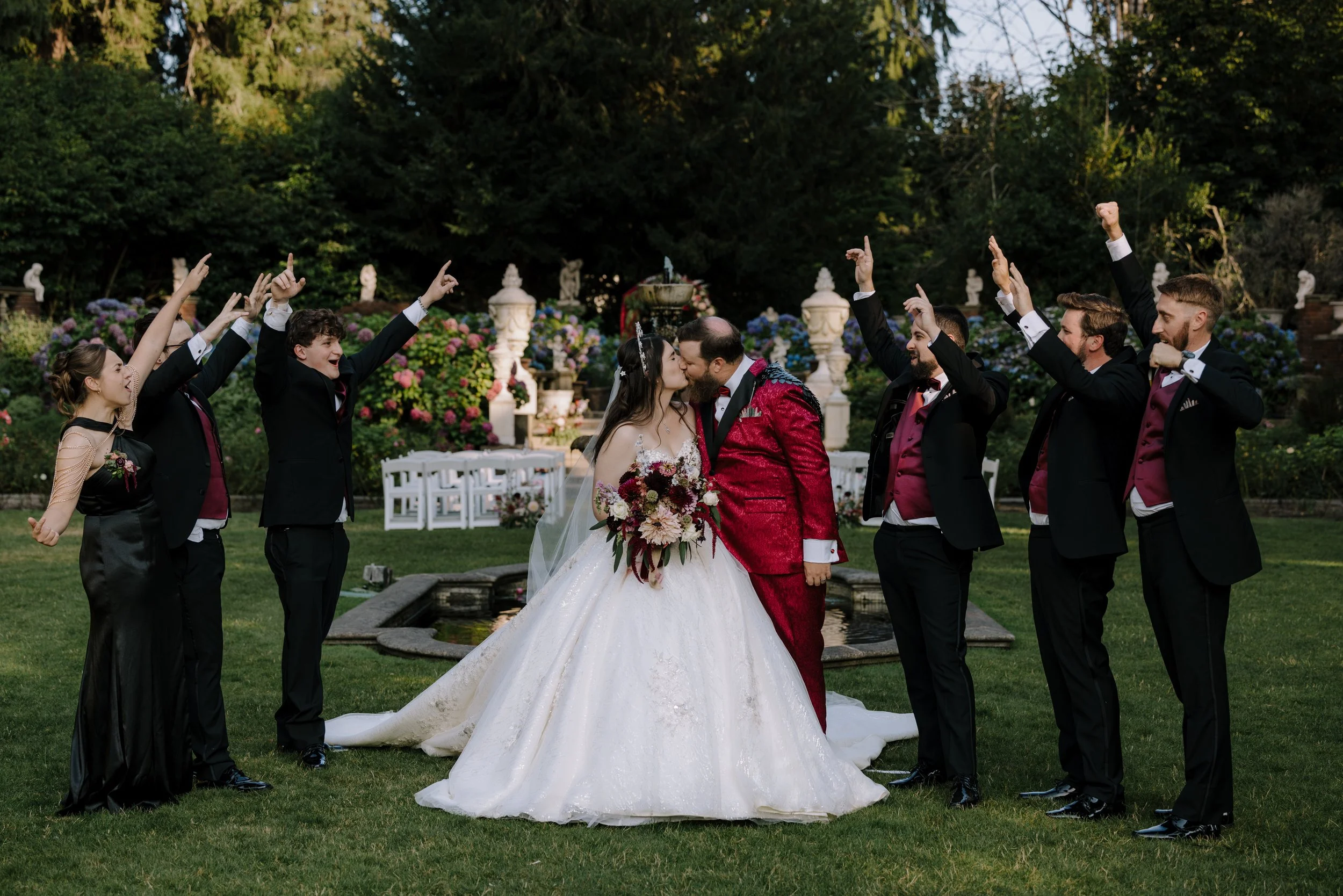 A wedding celebrating outdoors with a bride and groom kissing, surrounded by four men and one woman dressed in formal black suits with red vests and a woman in a black dress, all raising their hands in the air. Photo by Adventure Story Films