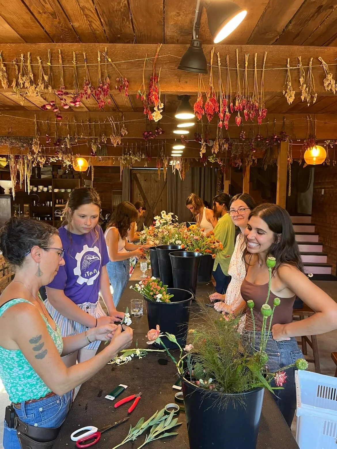 Group of women arranging flowers for a floral workshop in a rustic indoor setting with dried flowers hanging from the ceiling.