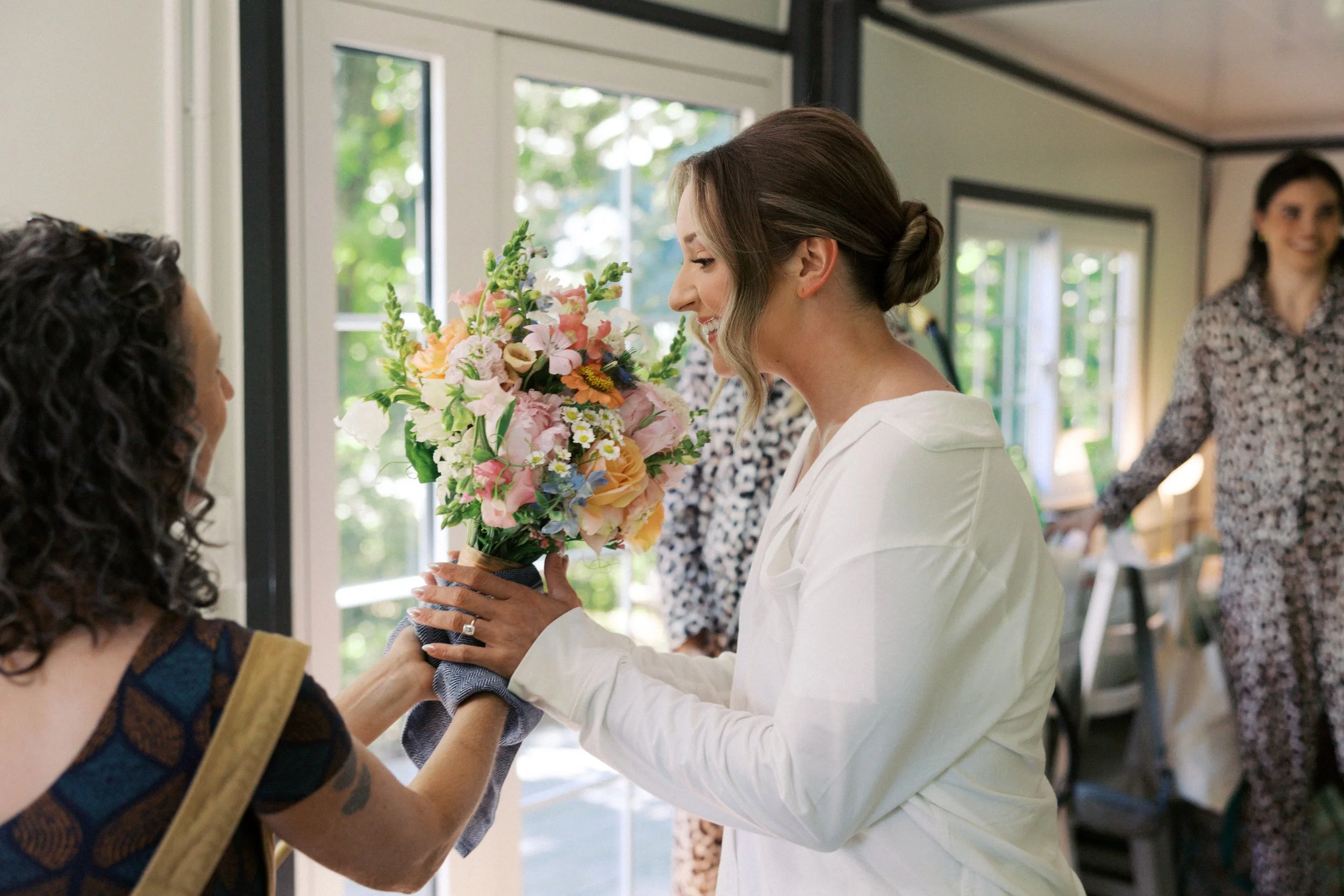 A woman in white receives a bouquet of flowers from another woman with curly hair and tattoos, inside a bright room with large windows and greenery outside. Photo by Here Today Photography.