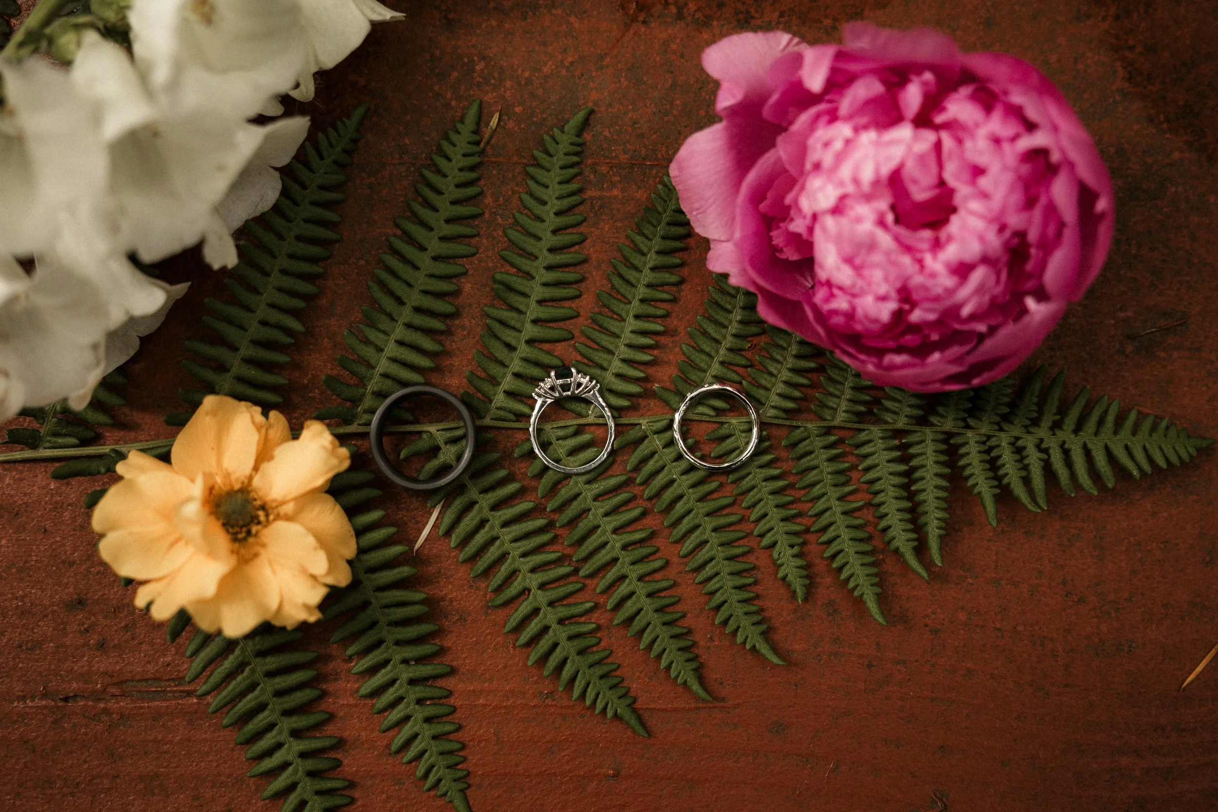 Three rings placed on the green fern leaves, surrounded by pink, yellow, and white flowers on a wooden surface.