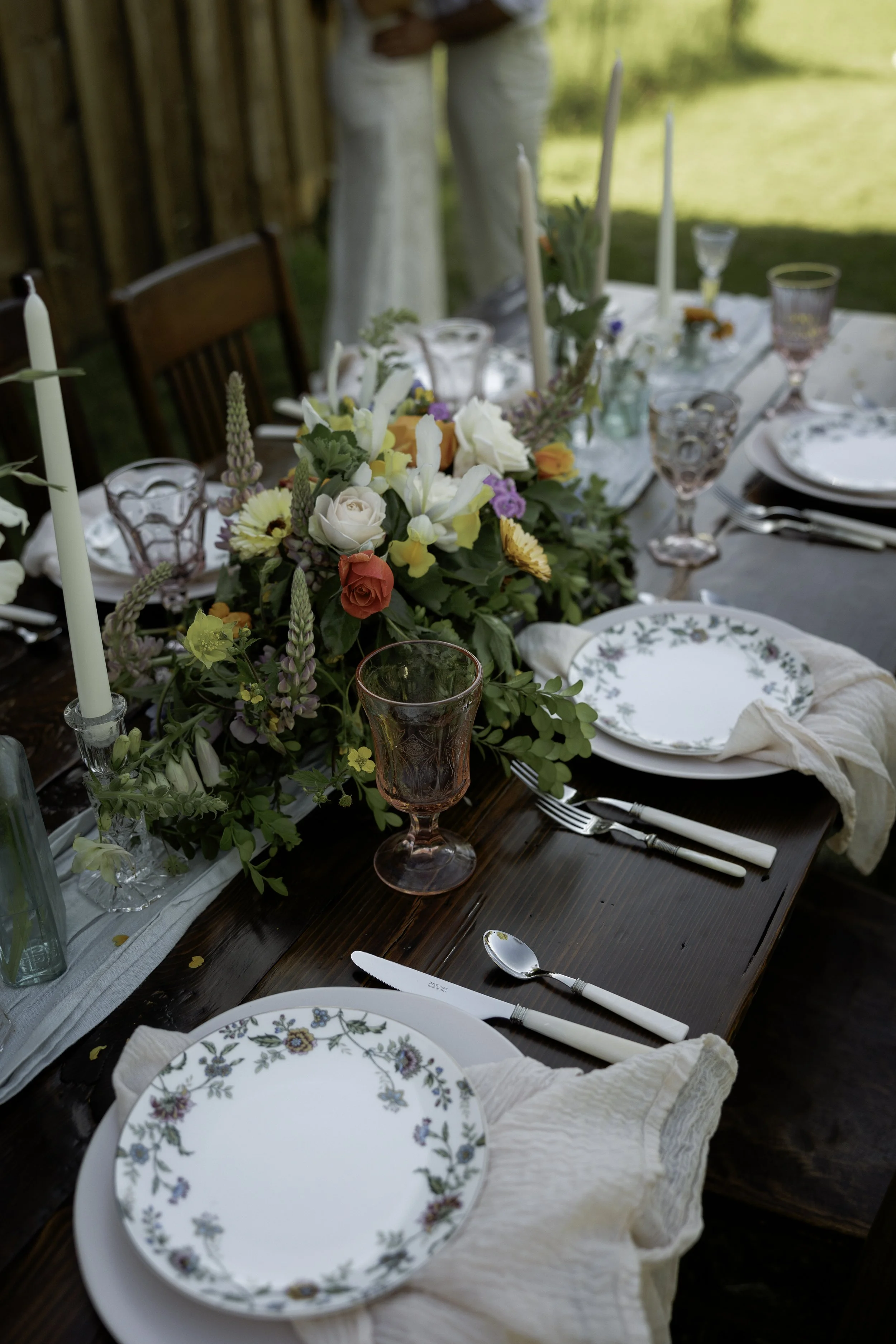 A beautifully set outdoor dining table with floral centerpiece, pink glassware, white plates with floral designs, white napkins, and silverware, surrounded by wooden chairs. Photo by Lauren Nunnally