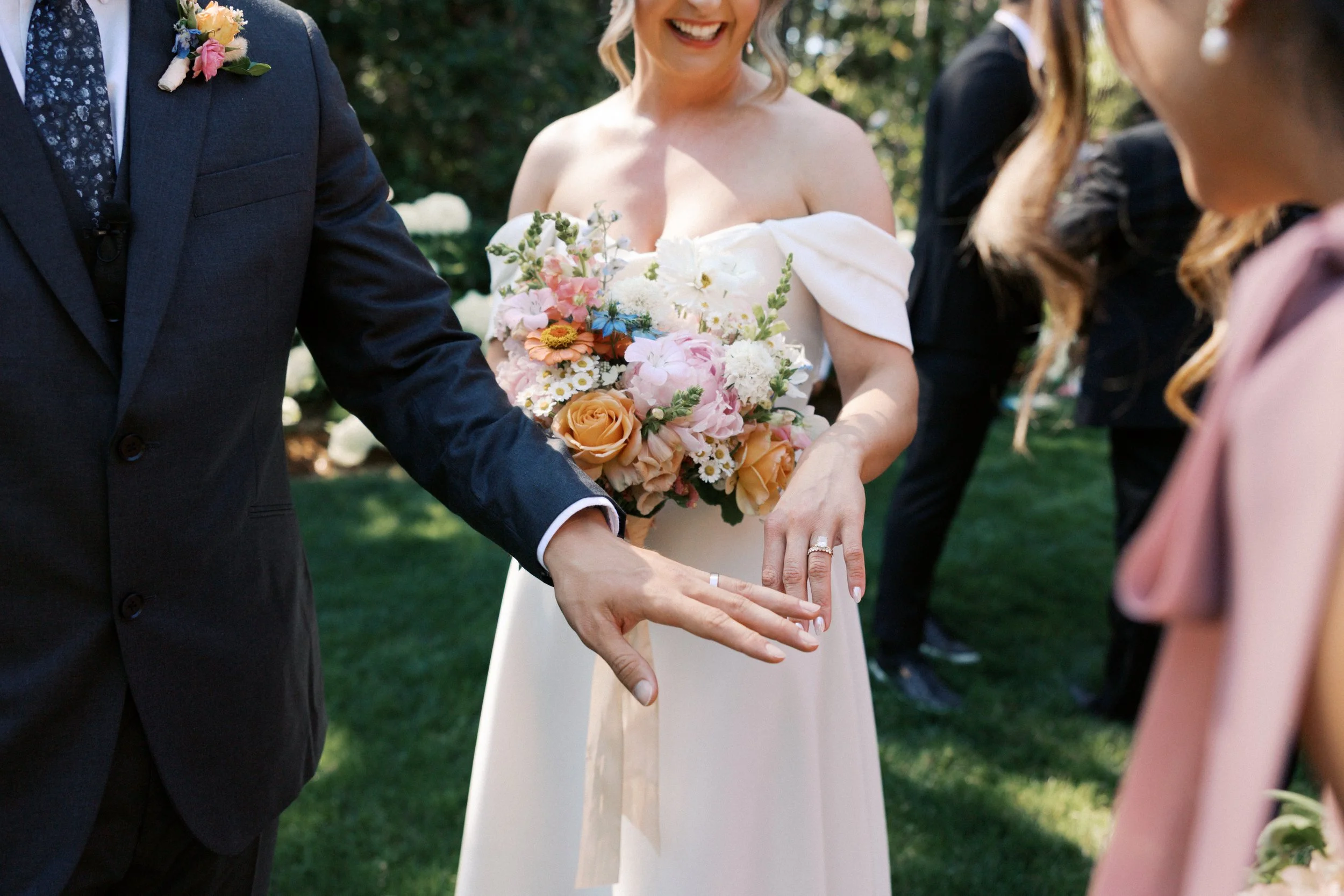 Wedding ceremony with bride holding a bouquet of colorful flowers, showing her wedding ring, and groom extending his hand, outdoors on grass with greenery in the background.

Photo by Here Today Photography
Venue: Rocky Hill Weddings & Events in Whit
