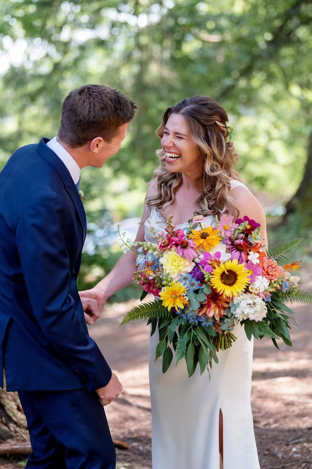A bride and groom laughing and holding hands in a wooded outdoor setting. The bride is holding a large colorful bouquet of flowers. Photo by Crystal Genes Photography