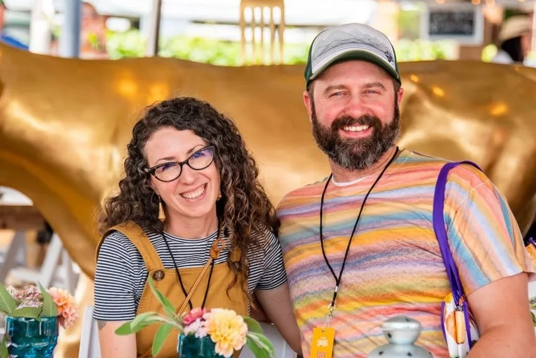 A photo of a smiling man and woman at a food and wine festival. There are flowers on the table in front of them. Photo by Whatever Photo PDX