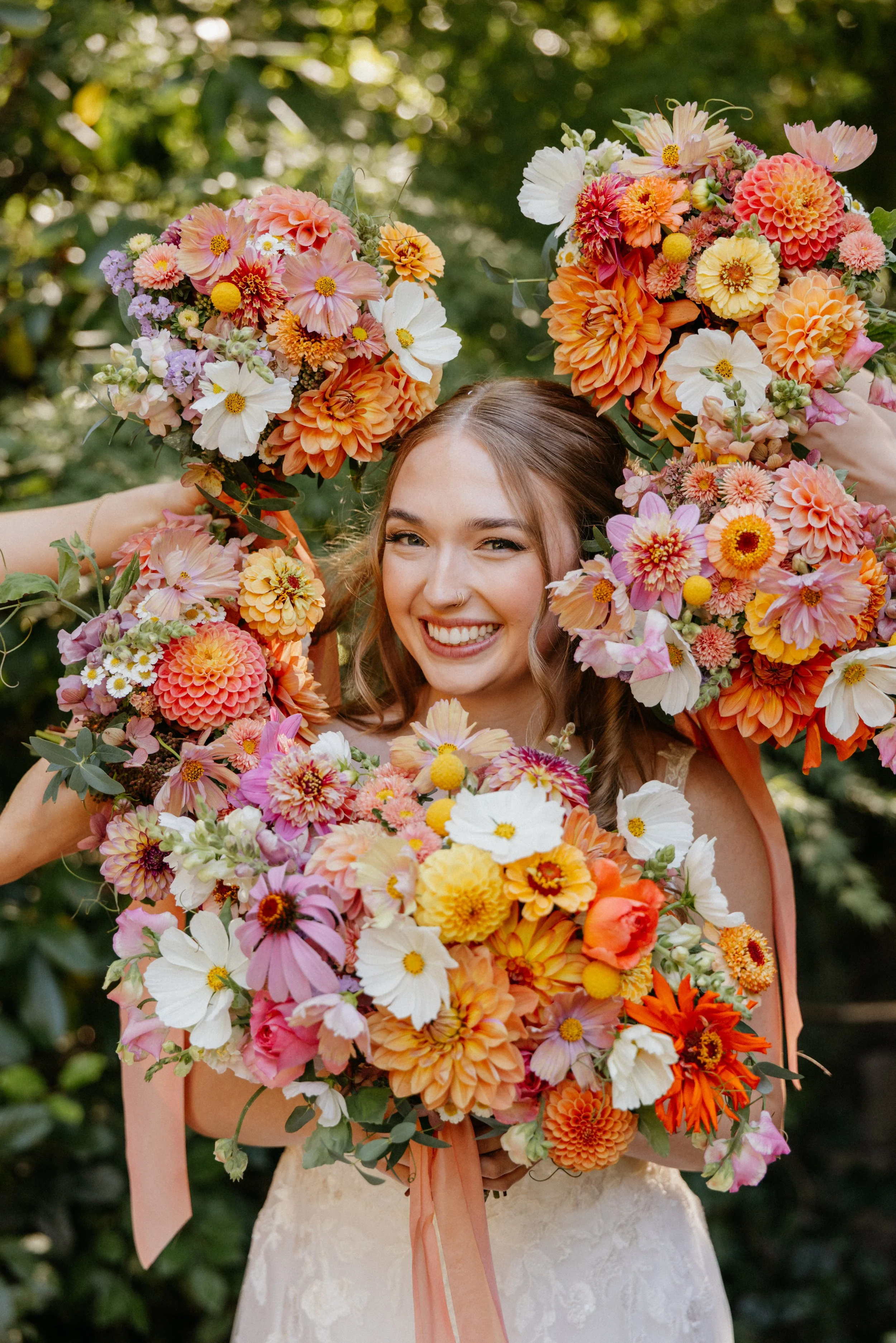 A young woman in a wedding dress holding large colorful bouquets of flowers with orange, pink, yellow, and white blossoms, smiling outdoors with greenery in the background.

Photo by Ivory Creek Photography
Venue: Summit Grove Lodge in Ridgefield, WA