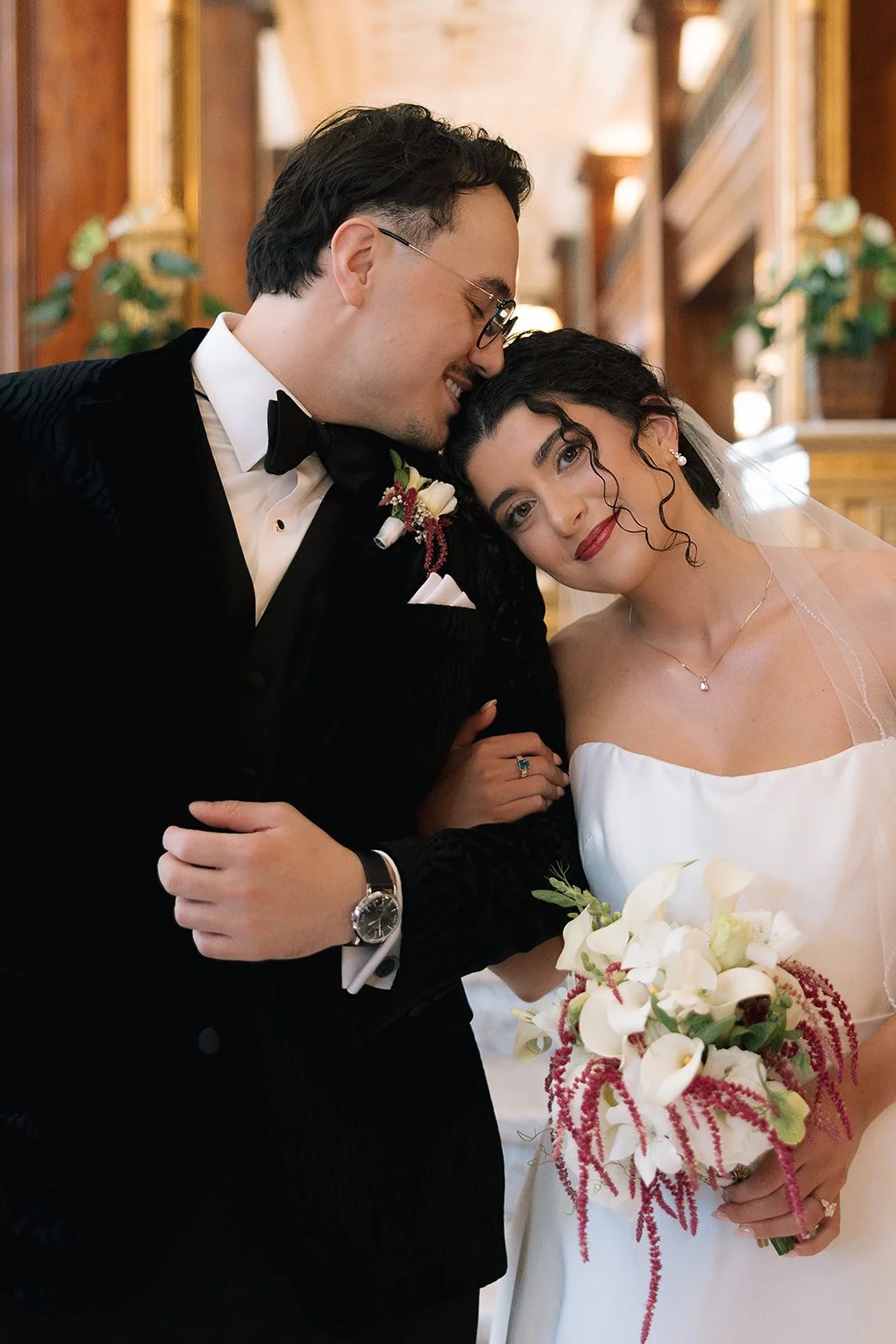 A  bride in a white wedding dress and holding a bouquet of calla lilies rests her head on her groom's shoulder. The groom is wearing a black tuxedo with a bow tie and a boutonniere. 

Photo: Mercedes Lou Photography
Venue: Benson Hotel in Portland, O