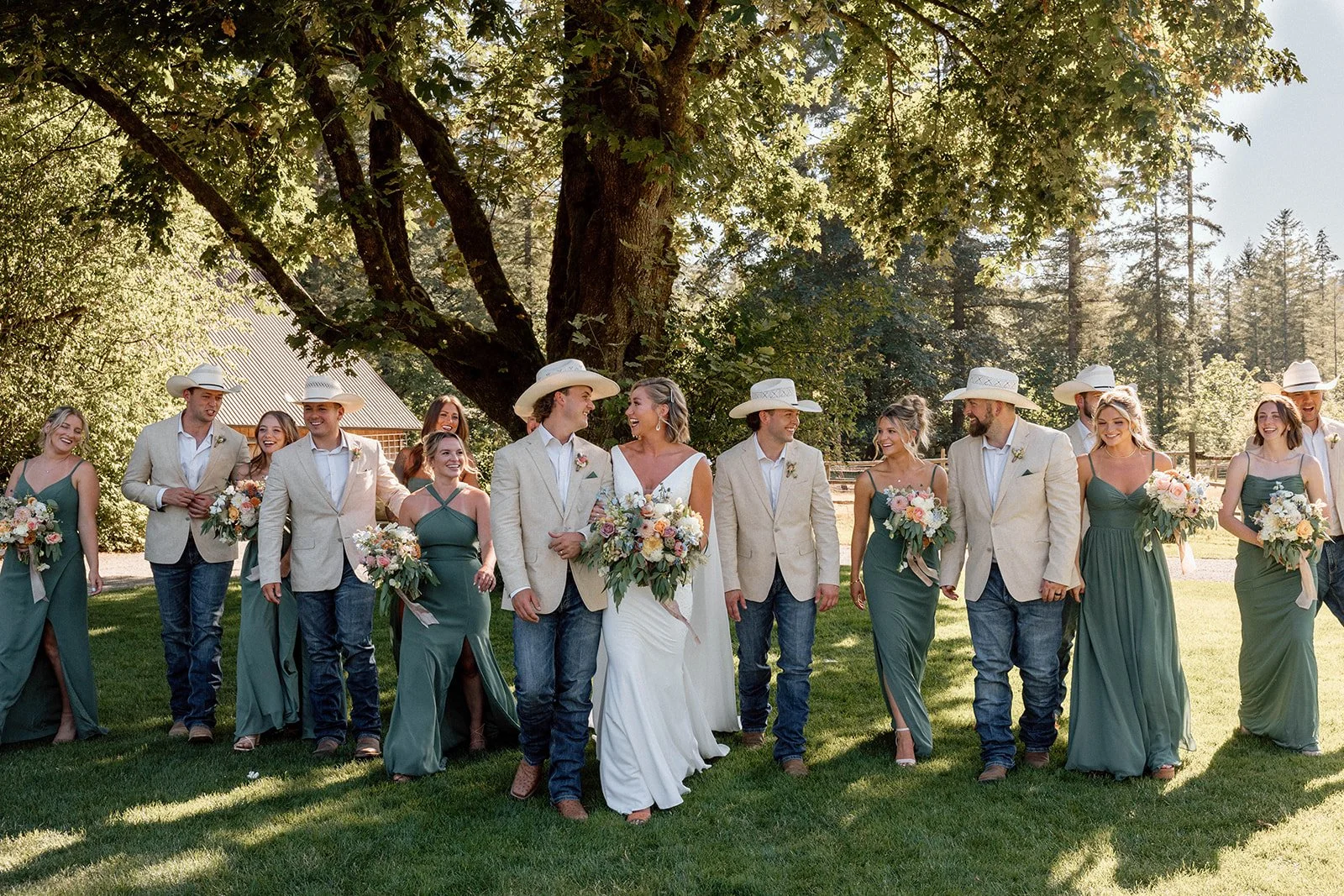 A wedding party walking outdoors under a large tree on a sunny day. The bride and groom are in the center, smiling and holding bouquets, with bridesmaids in green dresses and groomsmen in beige jackets and cowboy hats surrounding them.