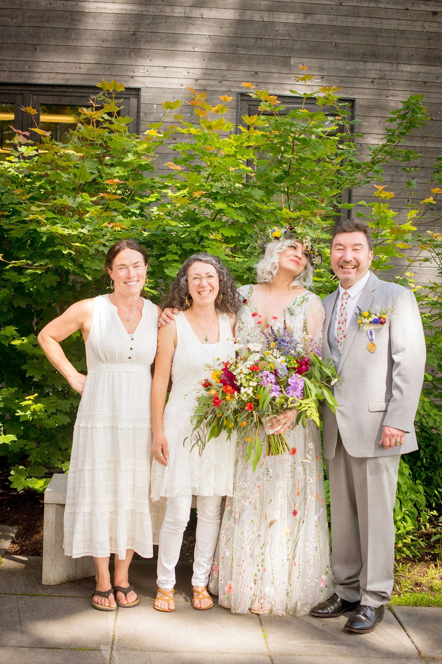 Group of five people, including a bride holding a large bouquet of flowers, standing outdoors in front of green bushes and a wooden wall.