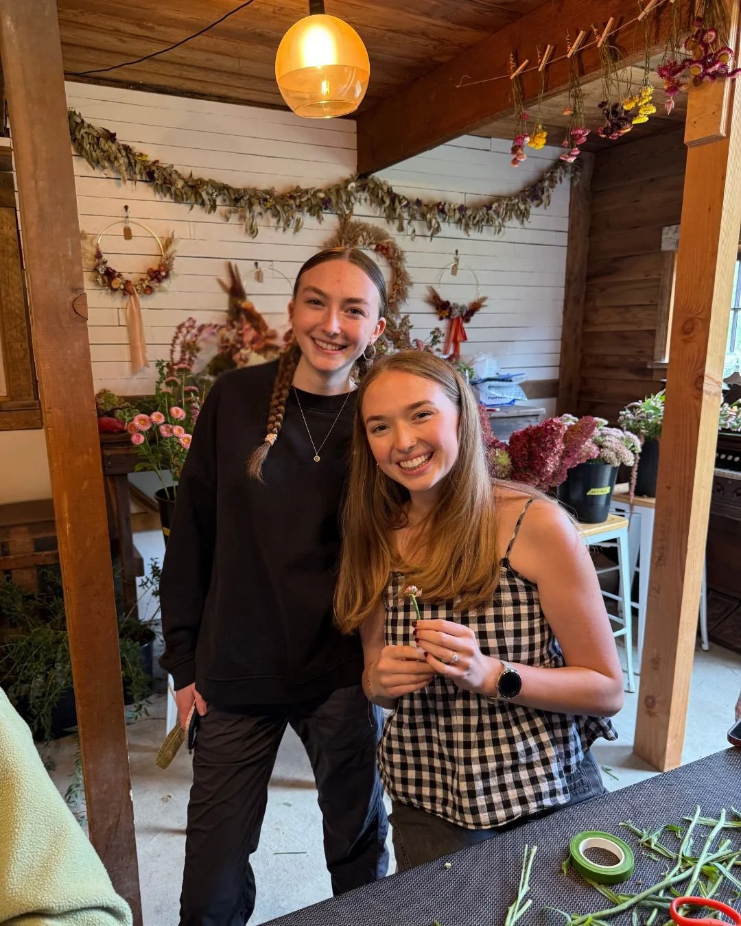 Two young women smiling indoors with a wooden and floral decor, one holding a small flower, surrounded by flowers and craft supplies.