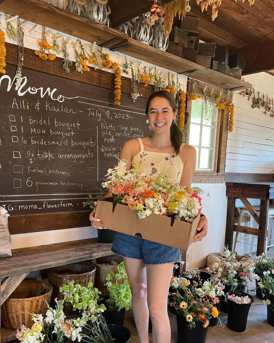 A young woman smiling and holding a box of mixed flowers inside a rustic flower shop with a chalkboard list of tasks, dried flowers hanging, and potted plants around.