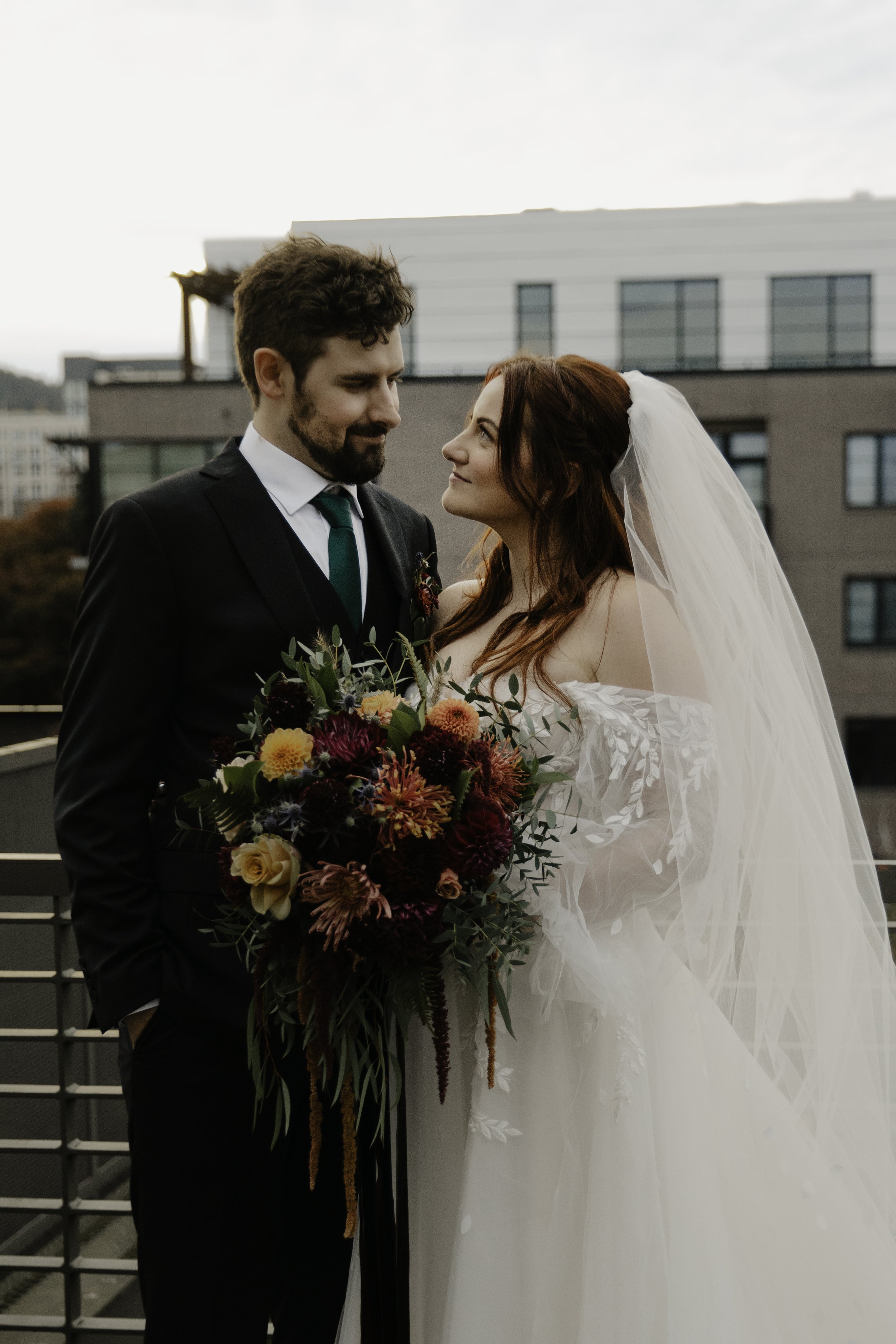 Bride and groom on a balcony, the bride holding a large, colorful bouquet, facing each other, the groom in a black suit with a green tie, and the bride in a white off-shoulder wedding dress with a veil, against an urban backdrop in Portland, Oregon.
