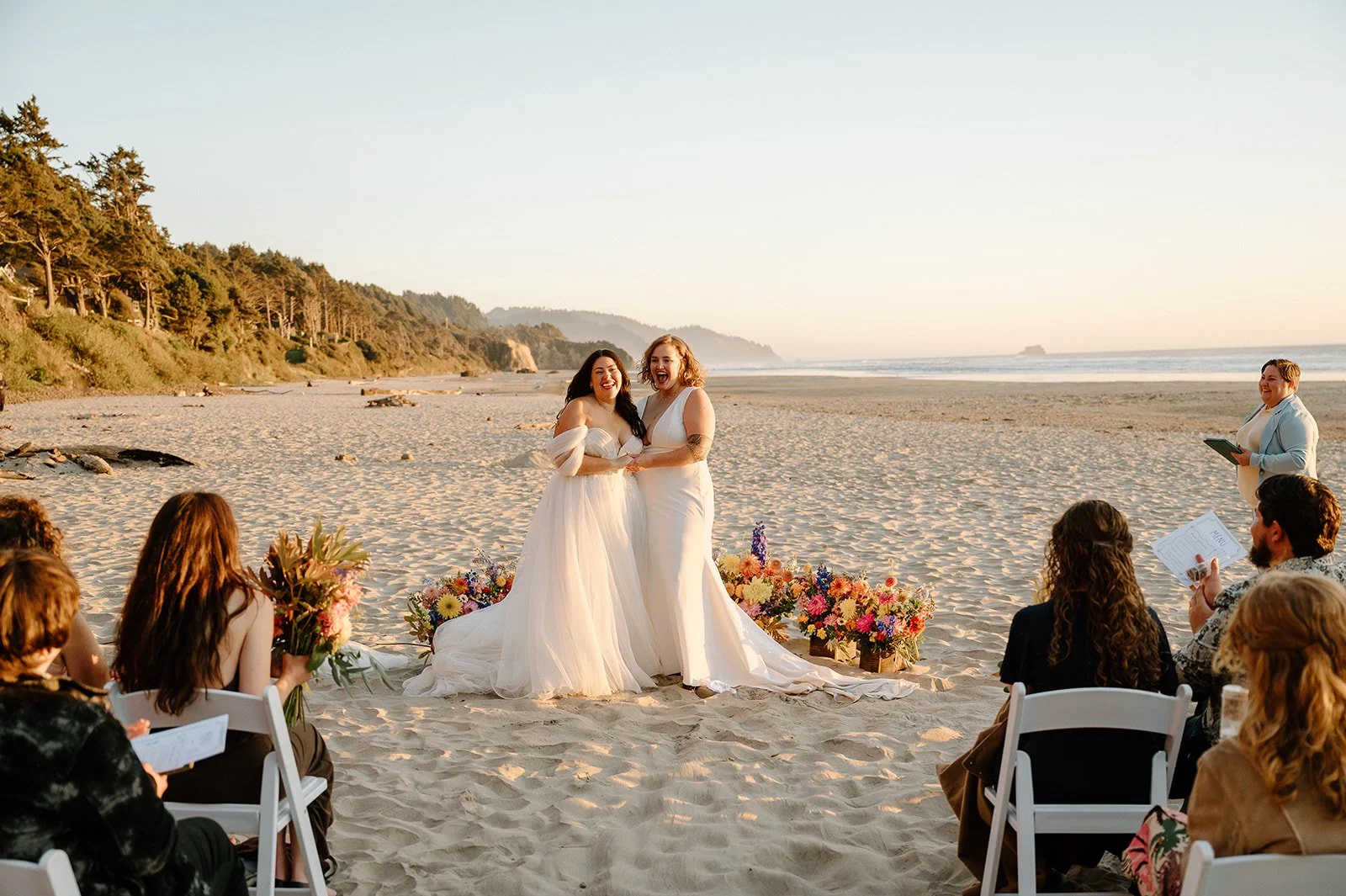 LGBTQ+ wedding ceremony on the beach with two brides, one in a strapless wedding dress and the other in a sleeveless gown, surrounded by friends and officiant, with colorful flowers on the sand, ocean, and cliffs in the background. Photo by Forgette