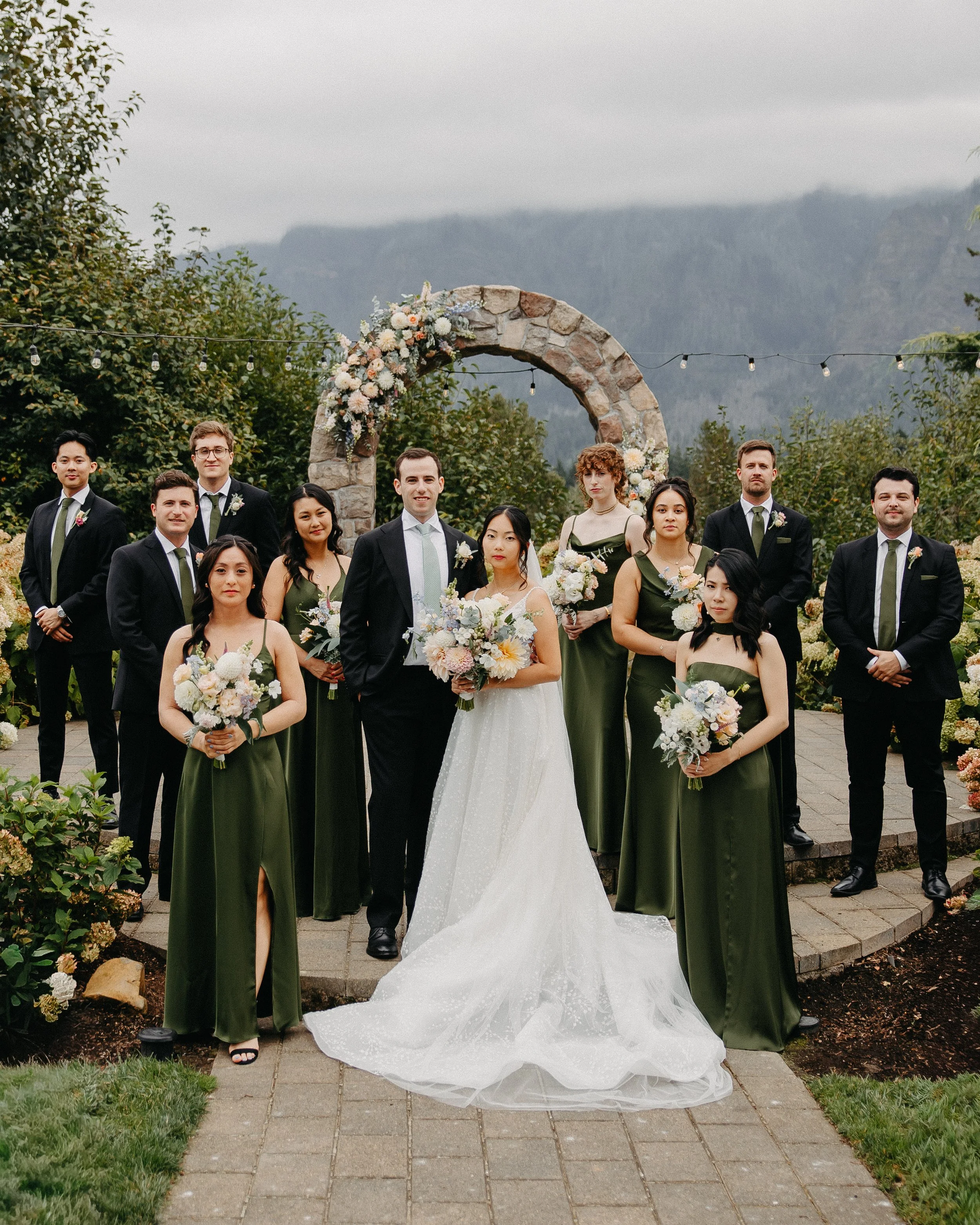 A wedding party with the bride, groom, and eight attendants standing outdoors in front of a stone arch decorated with flowers. The bride wears a white gown with a long train, and the bridesmaids wear green dresses. The groomsmen are in black suits wi