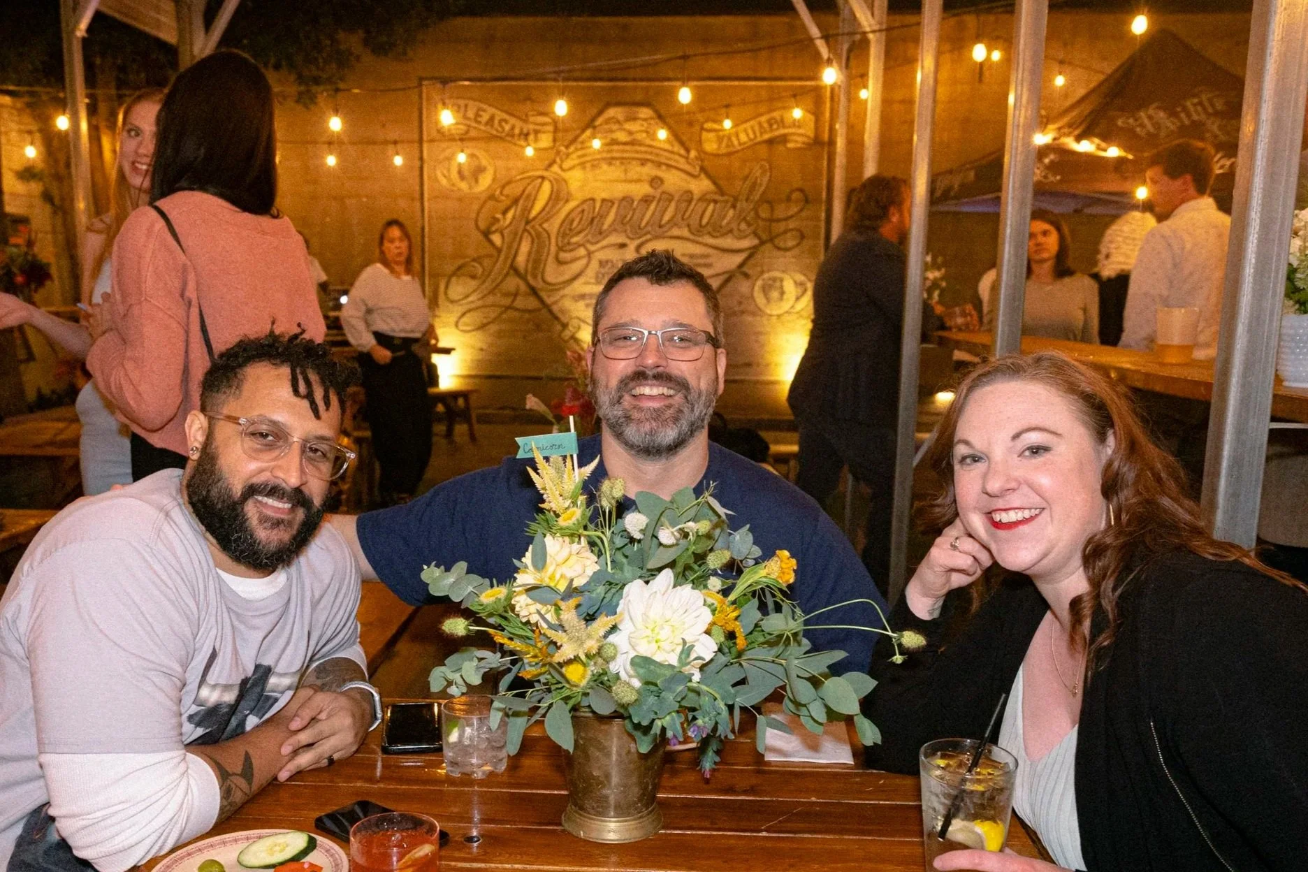 Three friends smiling at a table with a floral centerpiece at a social gathering or restaurant, with other people in the background. Photo by Whatever Photo PDX.