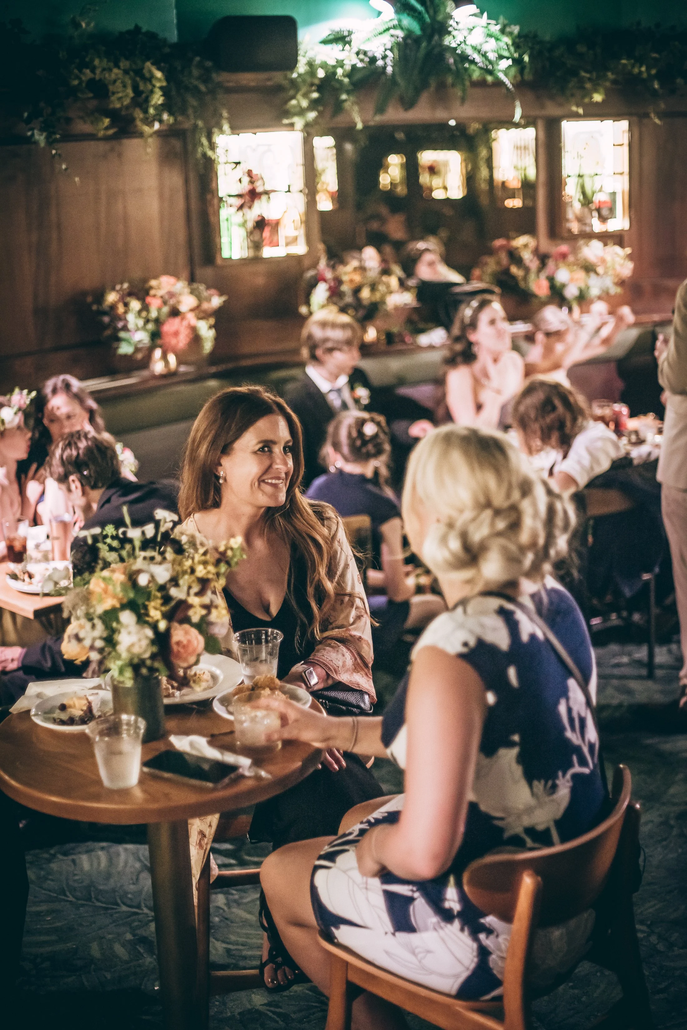 A woman with long hair smiling and holding a glass, sitting across from another woman with blonde hair, in a decorated dining area at a social event. Photo by Whatever Photo PDX.