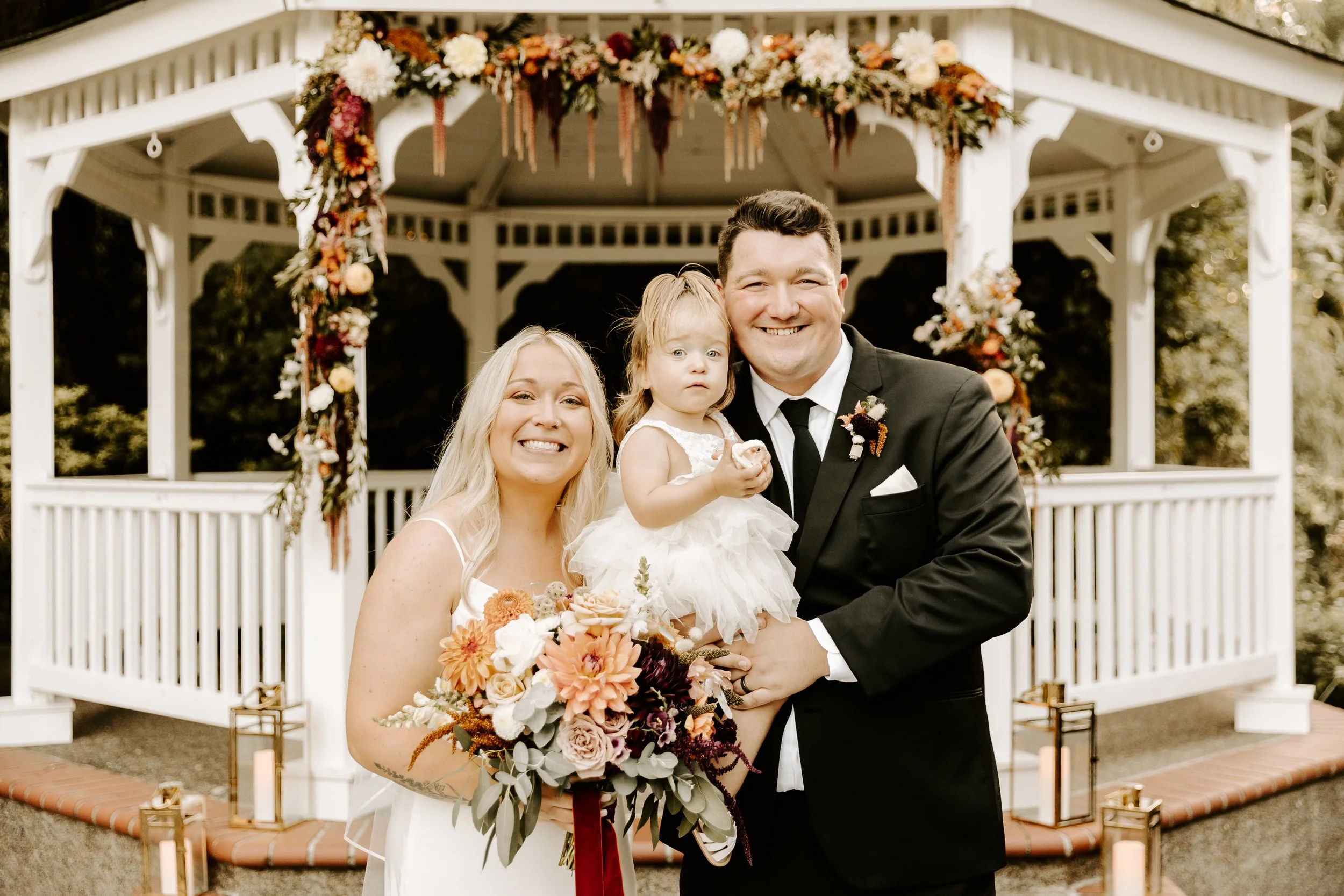 A newlywed couple with a young girl at a wedding outdoors in front of a decorated white gazebo, holding a bouquet and a cupcake.  Photo by B Sweet Imagery