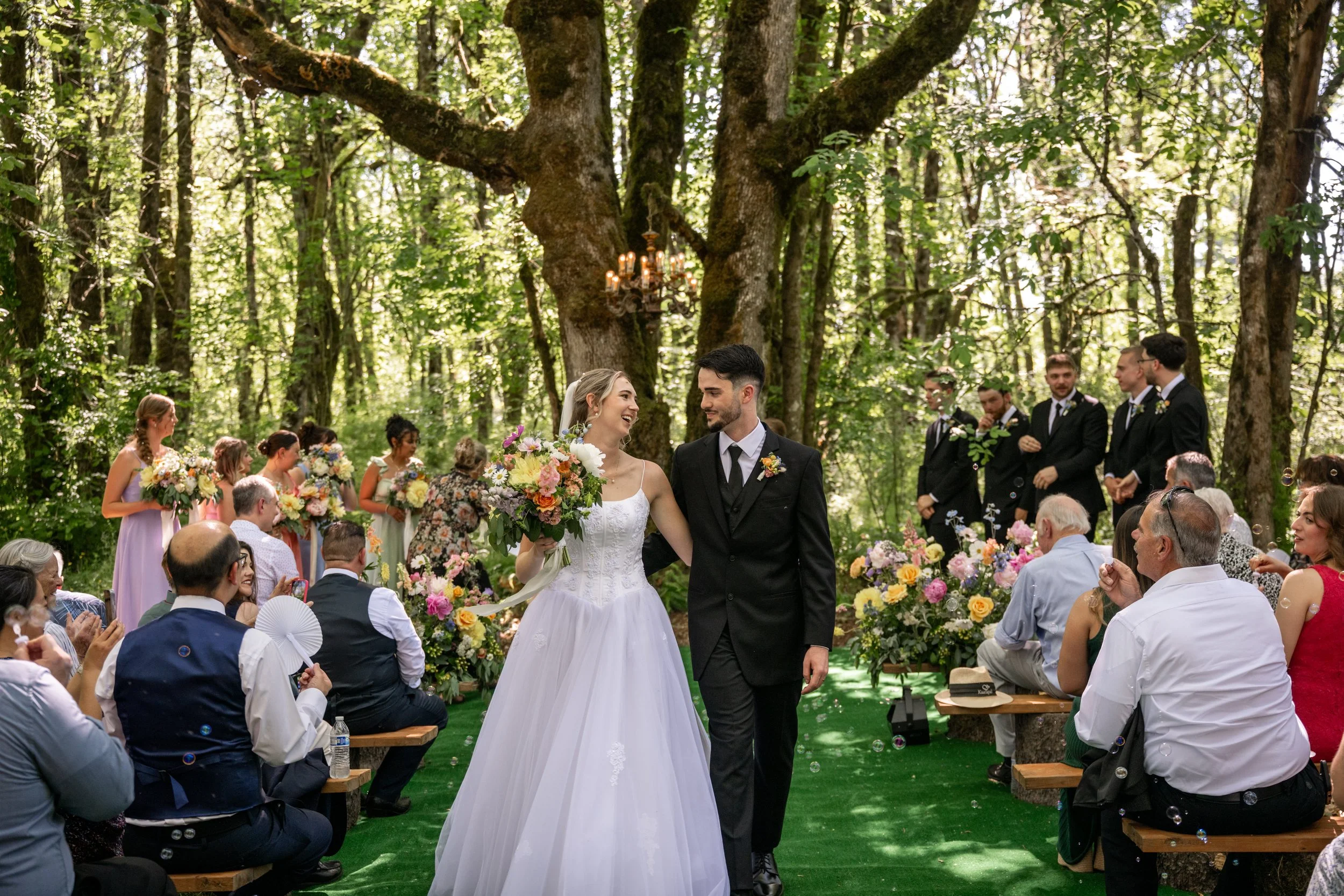 A bride and groom walking down the aisle at their outdoor wedding ceremony in a lush green forest, surrounded by seated guests, flower arrangements, and bridesmaids and groomsmen.

Photo by Red Letter Days
Venue: Vintage Gardens in Ridgefield, WA