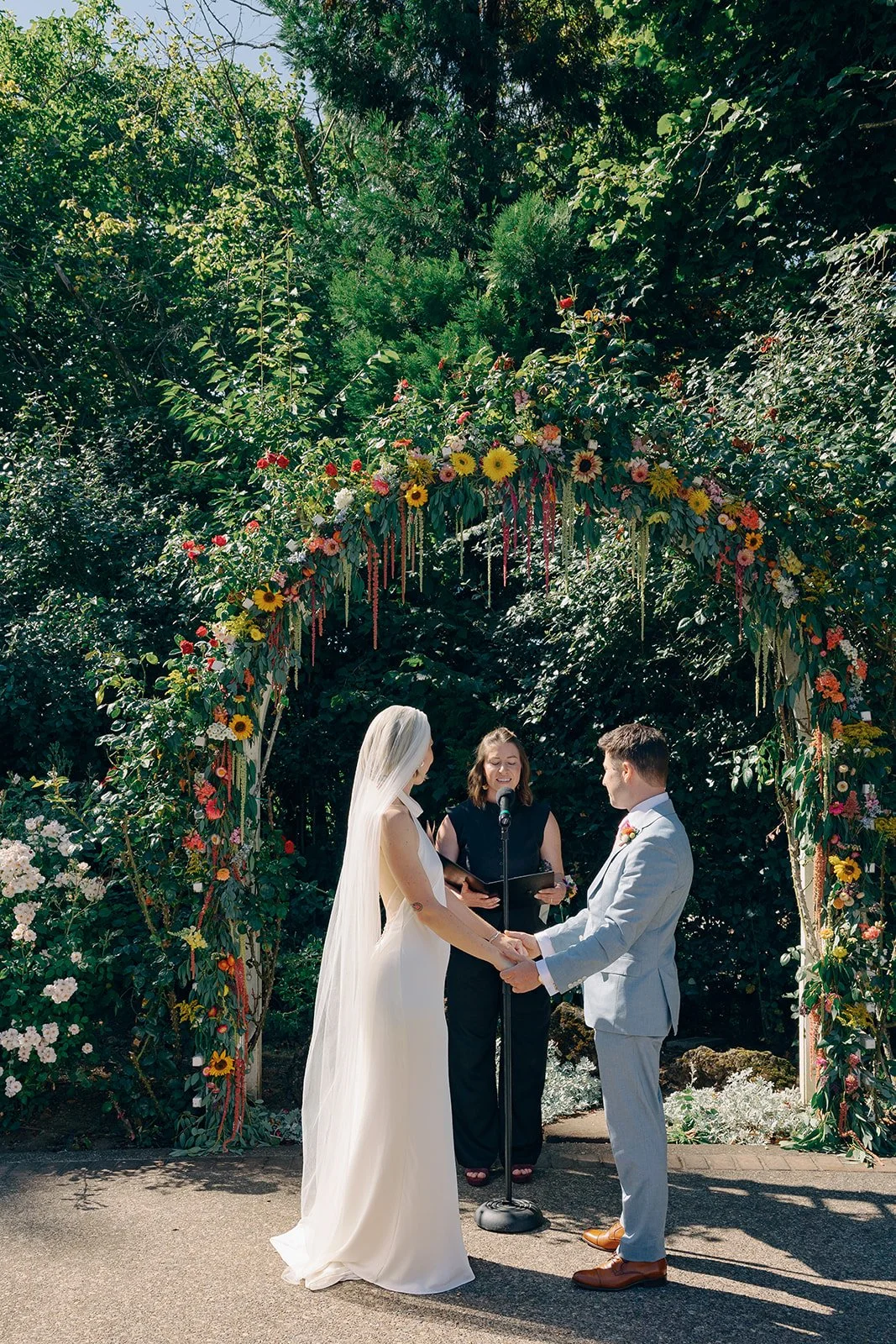 A couple getting married outdoors under a floral arch, holding hands and facing each other, with a woman officiating between them.