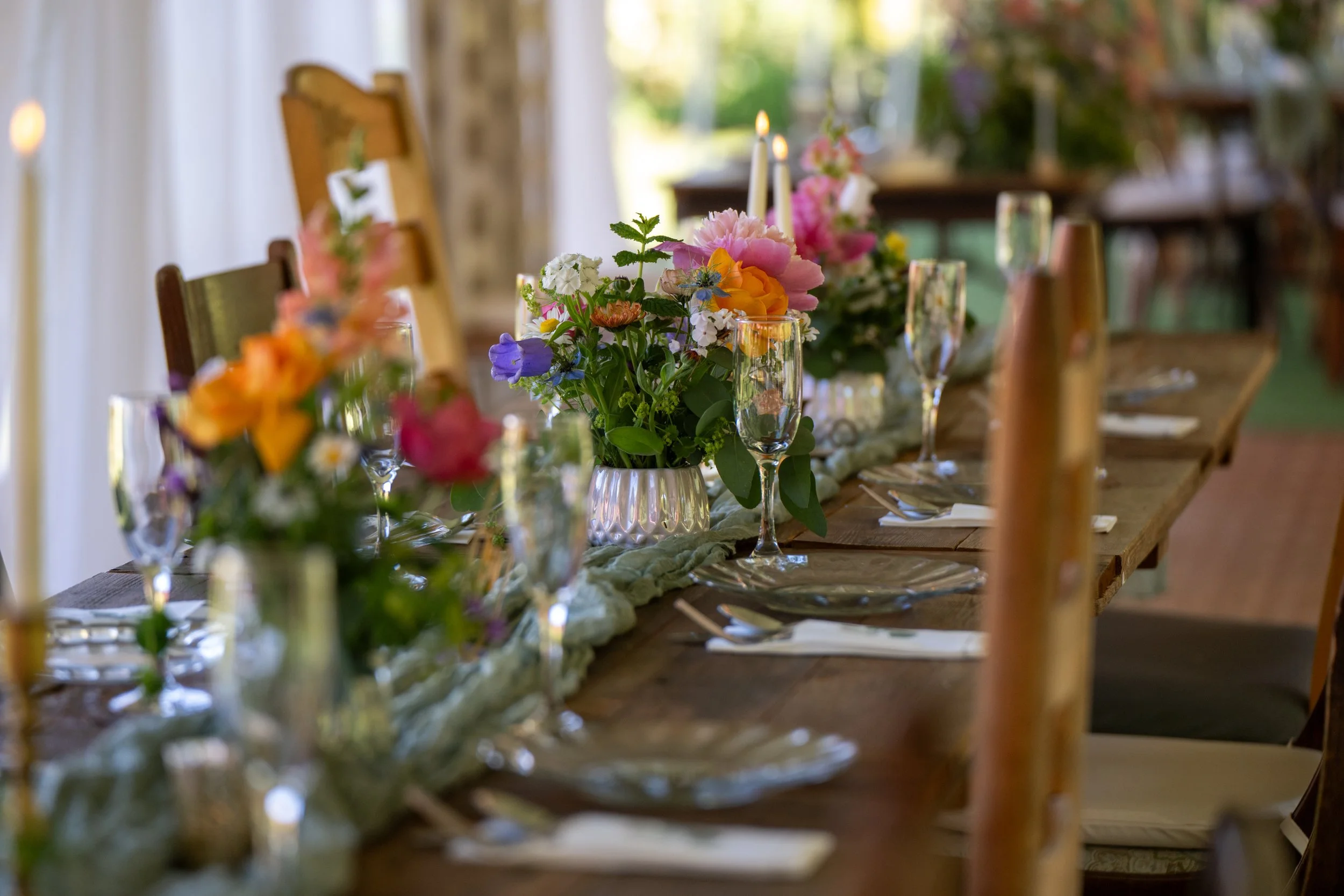 A beautifully decorated dining table with floral centerpieces, candles, and tableware, set outdoors with natural light. Photo by Red Letter Days