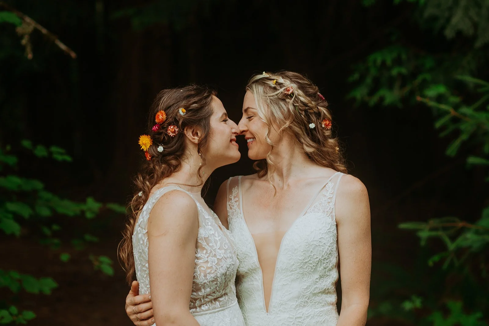 Two brides in white lace dresses standing close with noses touching in a forest setting, with flowers in their hair touching noses and smiling.

Photo by Olivia Louise Photography
Venue: Camp Cedarbrook in Vernonia, OR