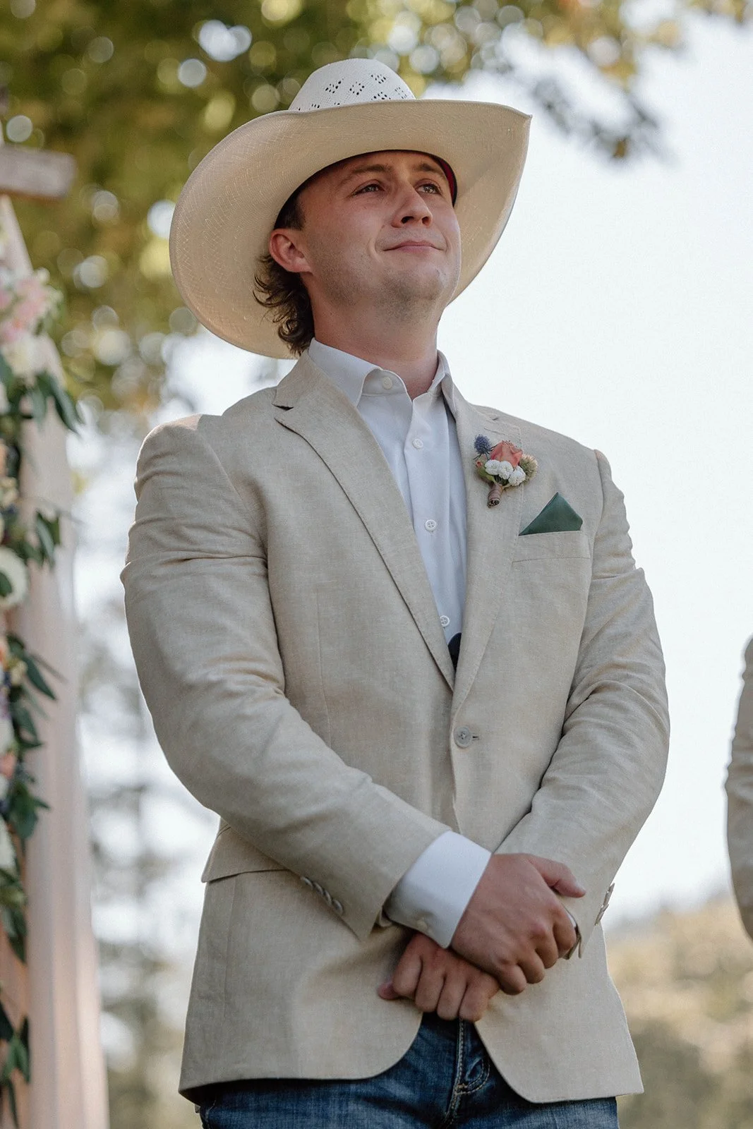 Man in beige blazer, white shirt, and a large white cowboy hat standing outdoors during a wedding ceremony.