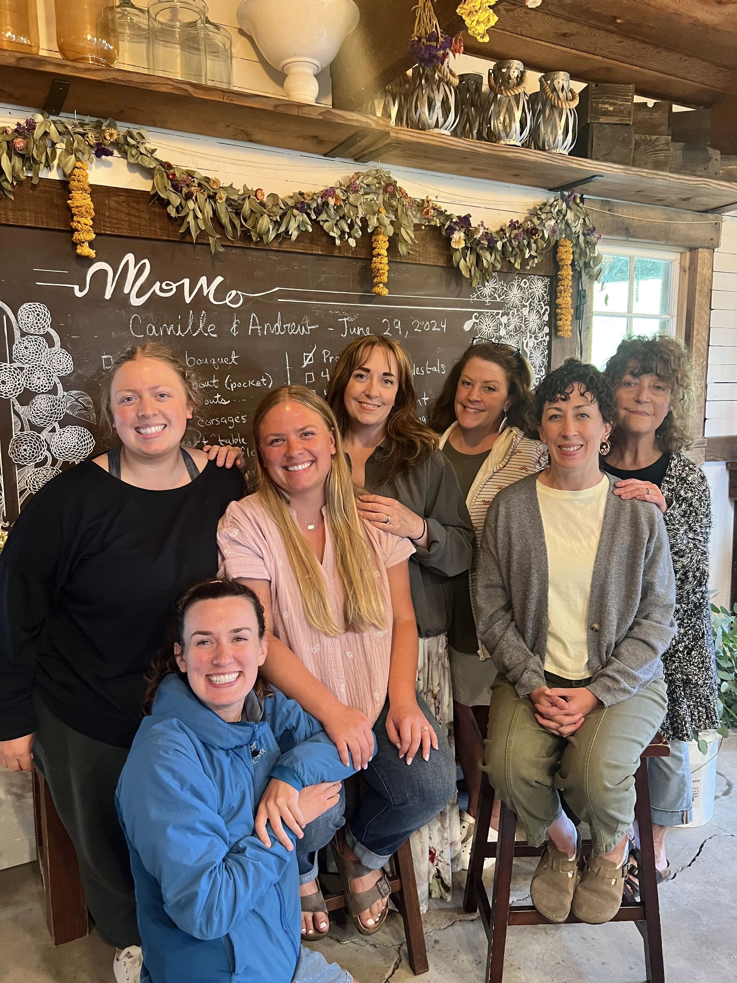 A group of seven women smiling and posing together in a cozy, rustic indoor space. There is a blackboard with handwritten notes and decorative floral garlands in the background.