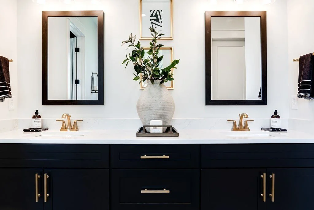 Double bathroom vanity with navy blue cabinets, white marble countertop, gold fixtures, two rectangular mirrors with black frames, a large vase with green foliage in the center, framed artwork above the vase, and black and white striped towels on each side.