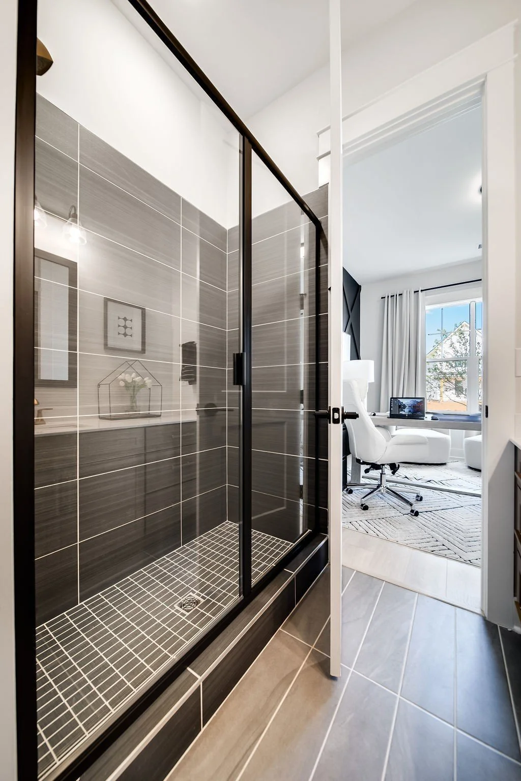 A modern bathroom with a glass shower enclosure and a view into a bright, white home office with a desk, computer, and large window with white curtains.