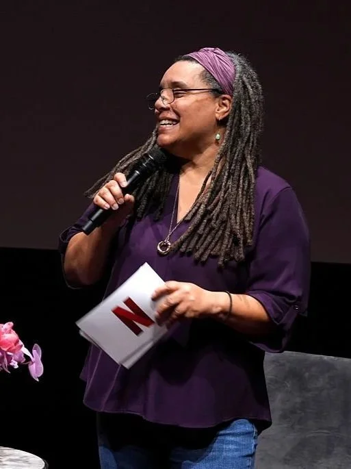 A woman with long dreadlocks wearing glasses, a purple headscarf, and a purple top, holding a microphone and a paper with a red 'N' on it, smiling on a stage.