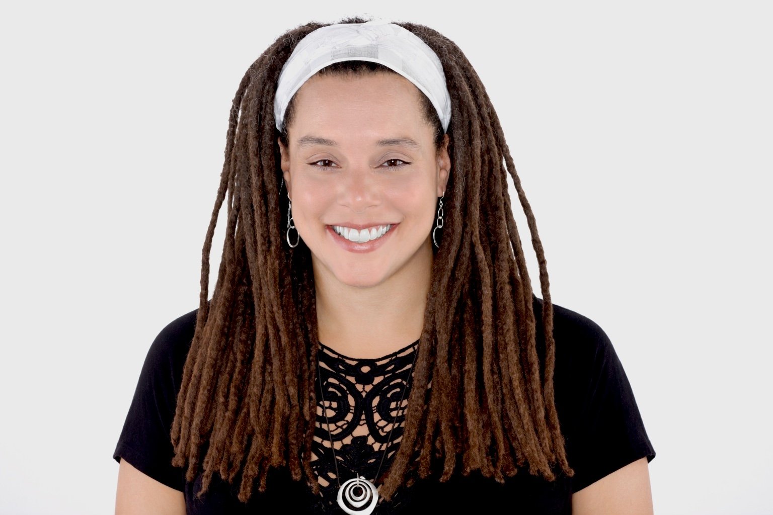 Woman with long dreadlocks, wearing a white headband, black top with lace detail, and silver jewelry, smiling against a plain gray background.
