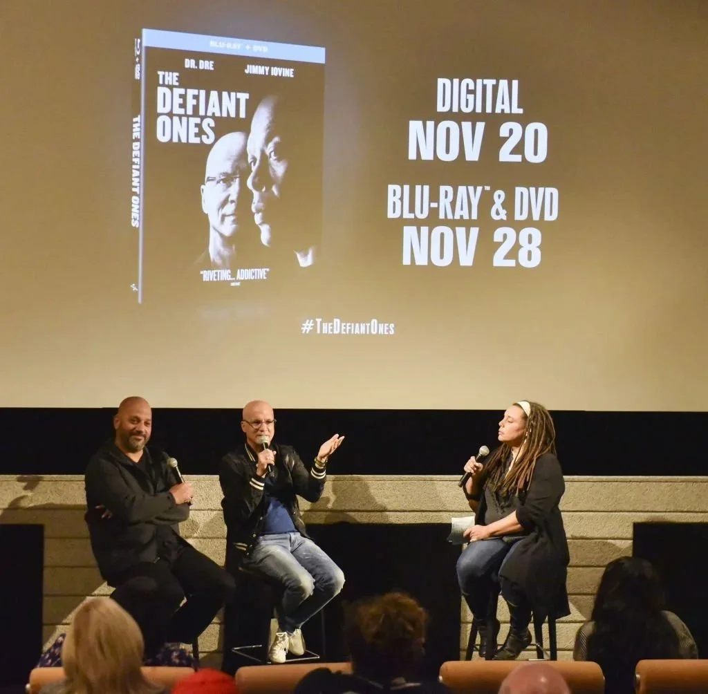Three people on stage in front of a large screen advertising the movie 'The Defiant Ones,' with release dates for digital and Blu-ray/DVD editions. The person on the left is holding a microphone and smiling, the middle person is speaking into a microphone, and the person on the right is listening and holding a microphone. An audience is sitting in front.