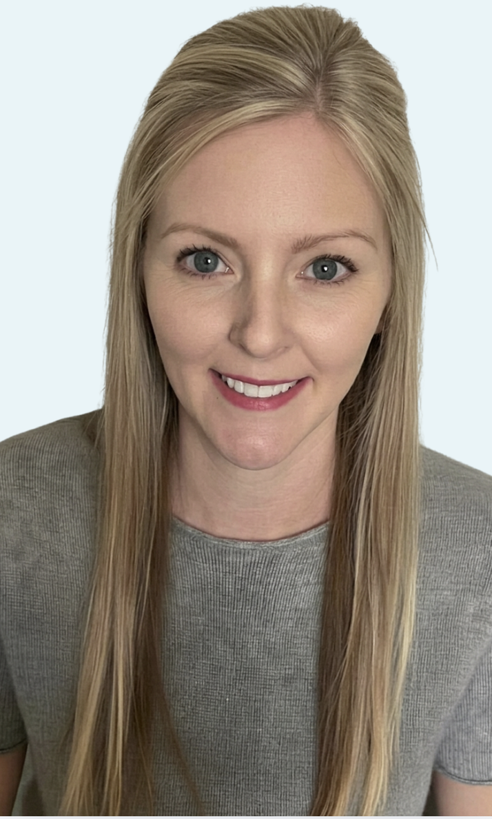 A woman with long blonde hair and green eyes smiling at the camera, wearing a light blue button-up shirt, in an indoor setting with blue walls and a door in the background.