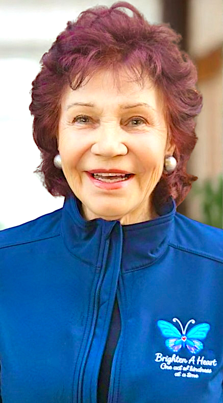 Smiling elderly woman with purple hair wearing pearl earrings and a blue jacket with a butterfly logo and text that reads 'Brighten A Heart, One act of kindness at a time.'