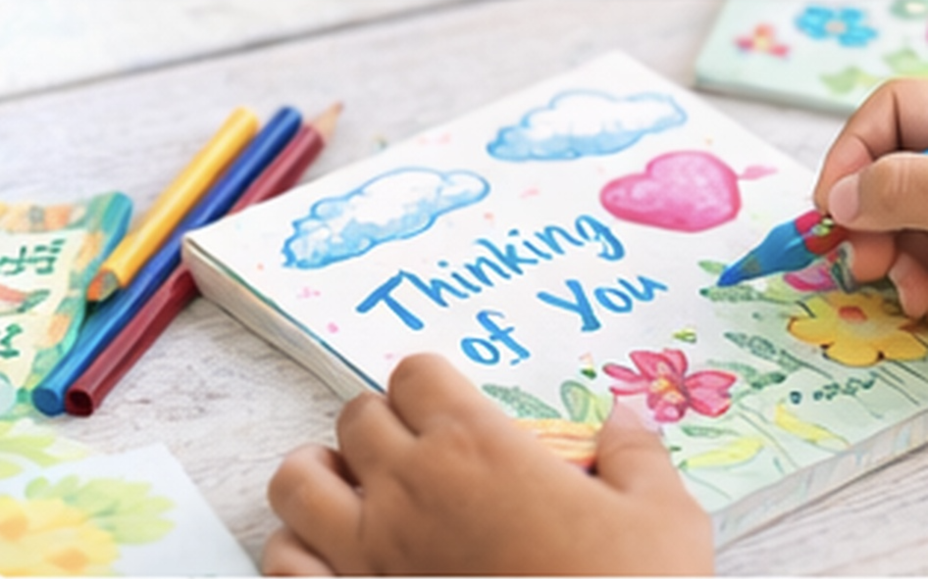 Hands drawing a colorful greeting card with the words "Thinking of You" and illustrations of clouds, hearts, and flowers.