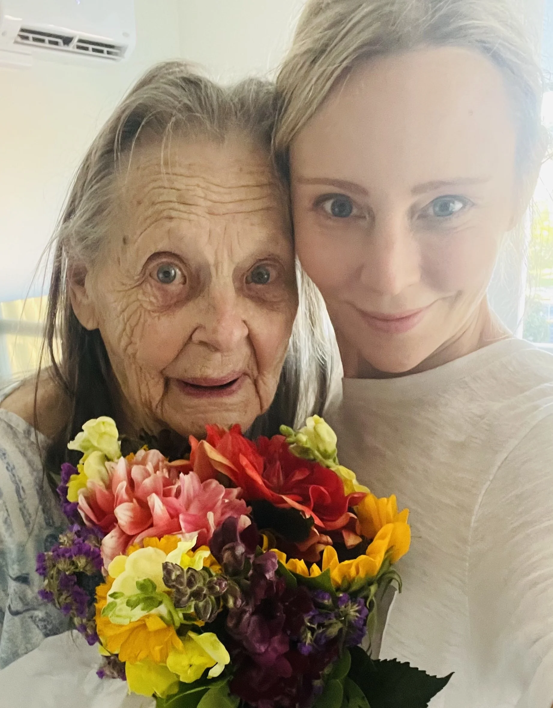 A young woman and an elderly woman take a selfie together, holding a vibrant bouquet of multicolored flowers, smiling with a background of a light-colored room and window