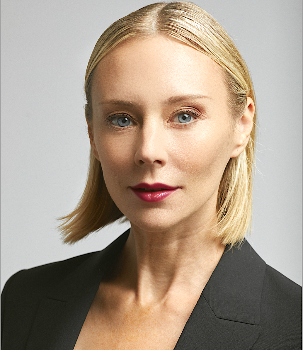 A professional woman with blonde hair, blue eyes, wearing a black blazer and red lipstick, looking directly at the camera with a neutral expression.