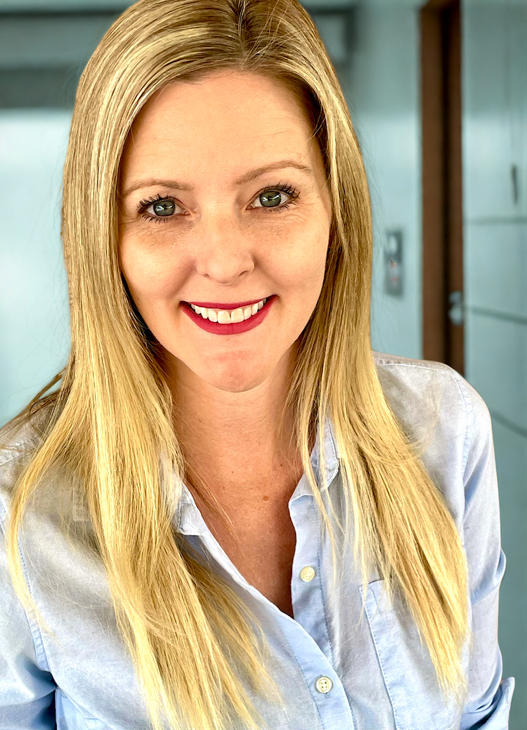 A woman with long blonde hair and green eyes smiling at the camera, wearing a light blue button-up shirt, in an indoor setting with blue walls and a door in the background.
