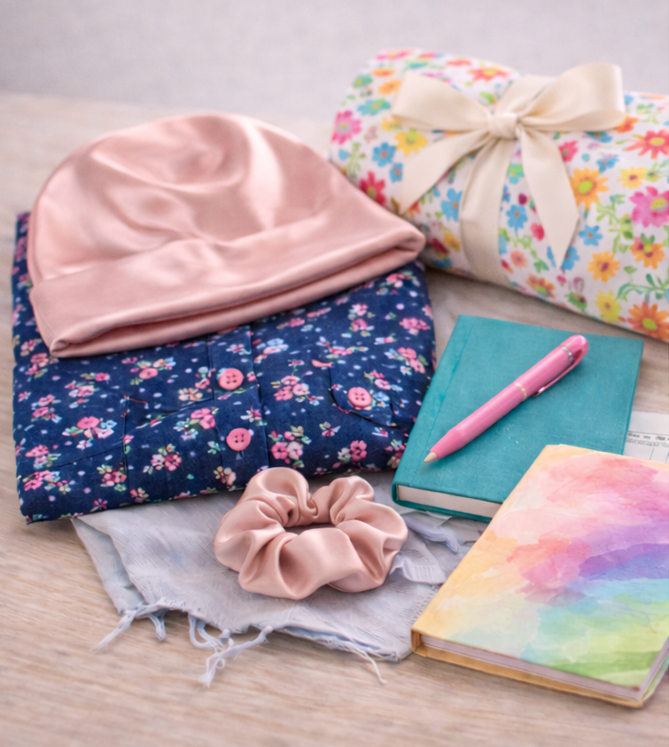 A collection of women's accessories and stationery items on a wooden surface, including a pink satin headscarf, a floral quilted skirt with pink buttons, a pink scrunchie, a teal notebook with a pink pen, and two colorful notebooks wrapped with a floral gift ribbon.
