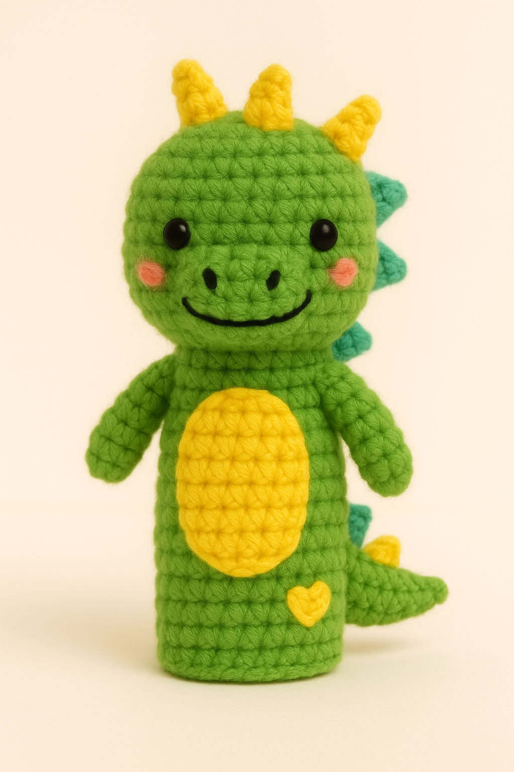 A crocheted plush toy resembling a cute green dinosaur with yellow horns, a yellow belly, and a small yellow heart on its lower body. The dinosaur has black eyes, pink cheeks, and a smiling face.