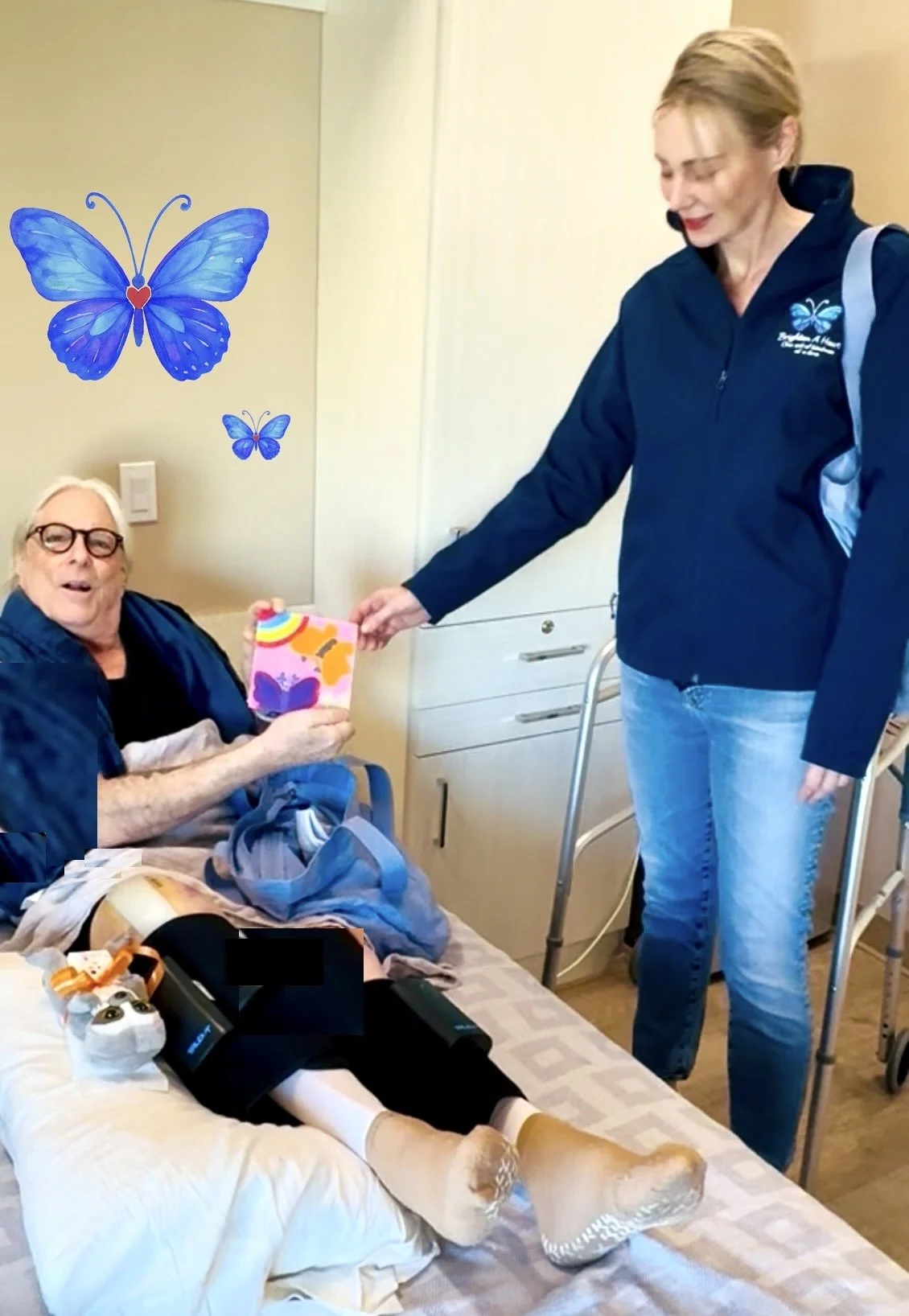 An elderly man with a bandaged leg in a hospital bed, smiling and receiving a colorful handmade card from a woman standing beside him. The woman has short blond hair, is wearing a blue jacket, and is holding a walker nearby. There are blue butterfly decorations on the wall.