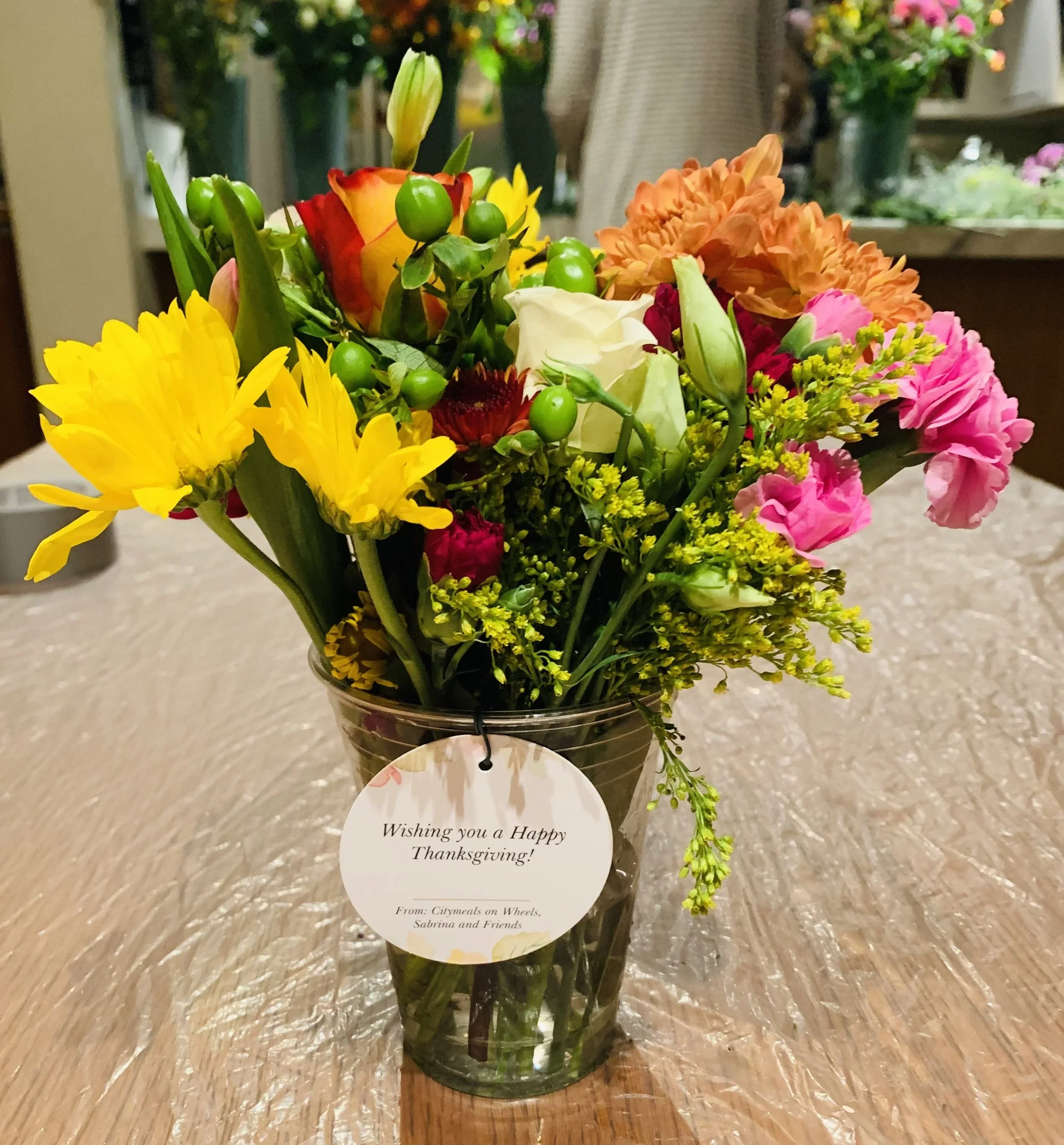Colorful mixed flower bouquet in a glass vase on a table, with a Thanksgiving message tag attached.