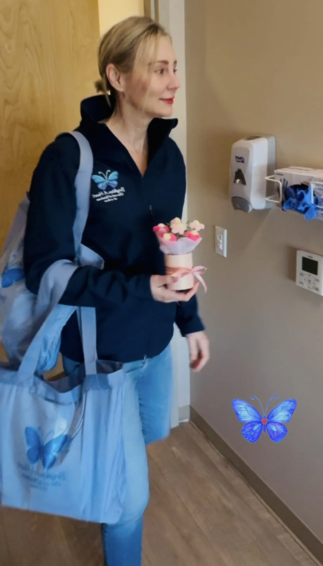 A woman standing in a hospital hallway holding a pink box of flowers, wearing a 'Daniel' embroidered jacket, with a blue butterfly sticker on her nylon bag and a butterfly sticker on the wall.