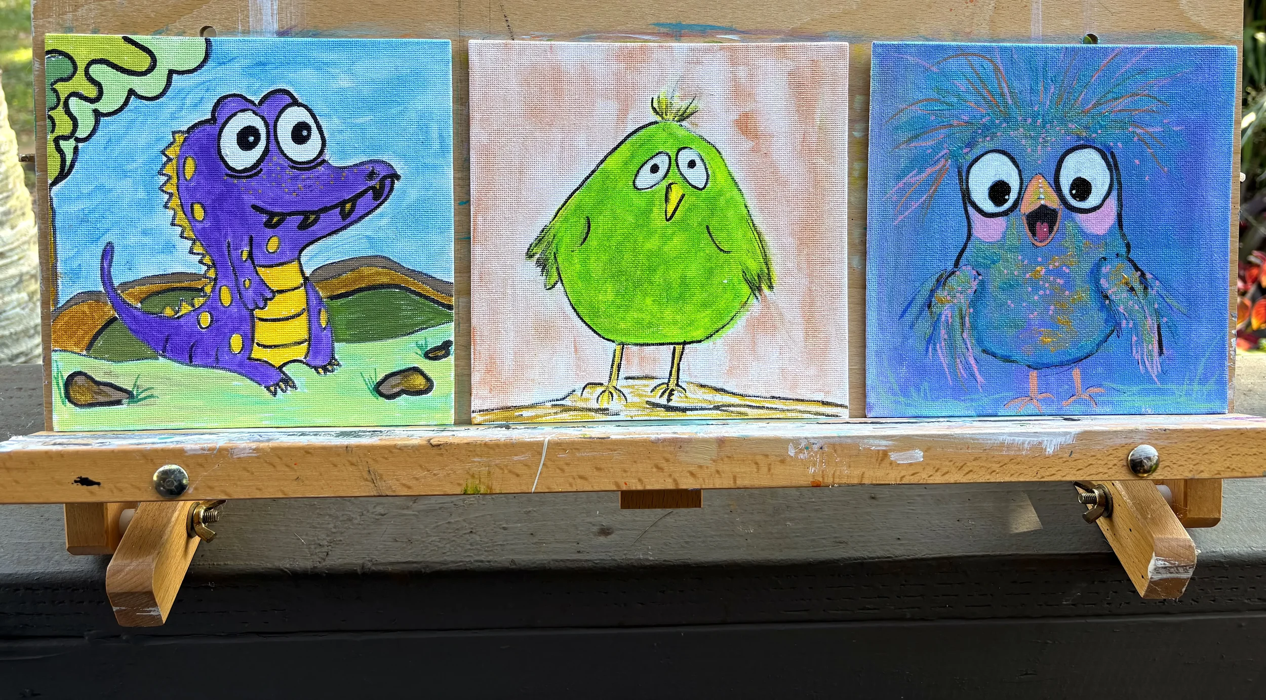 Three colorful cartoon style paintings of animals on small canvases, featuring a purple dinosaur, a green bird, and a blue owl, leaning on a wooden display in an outdoor setting.