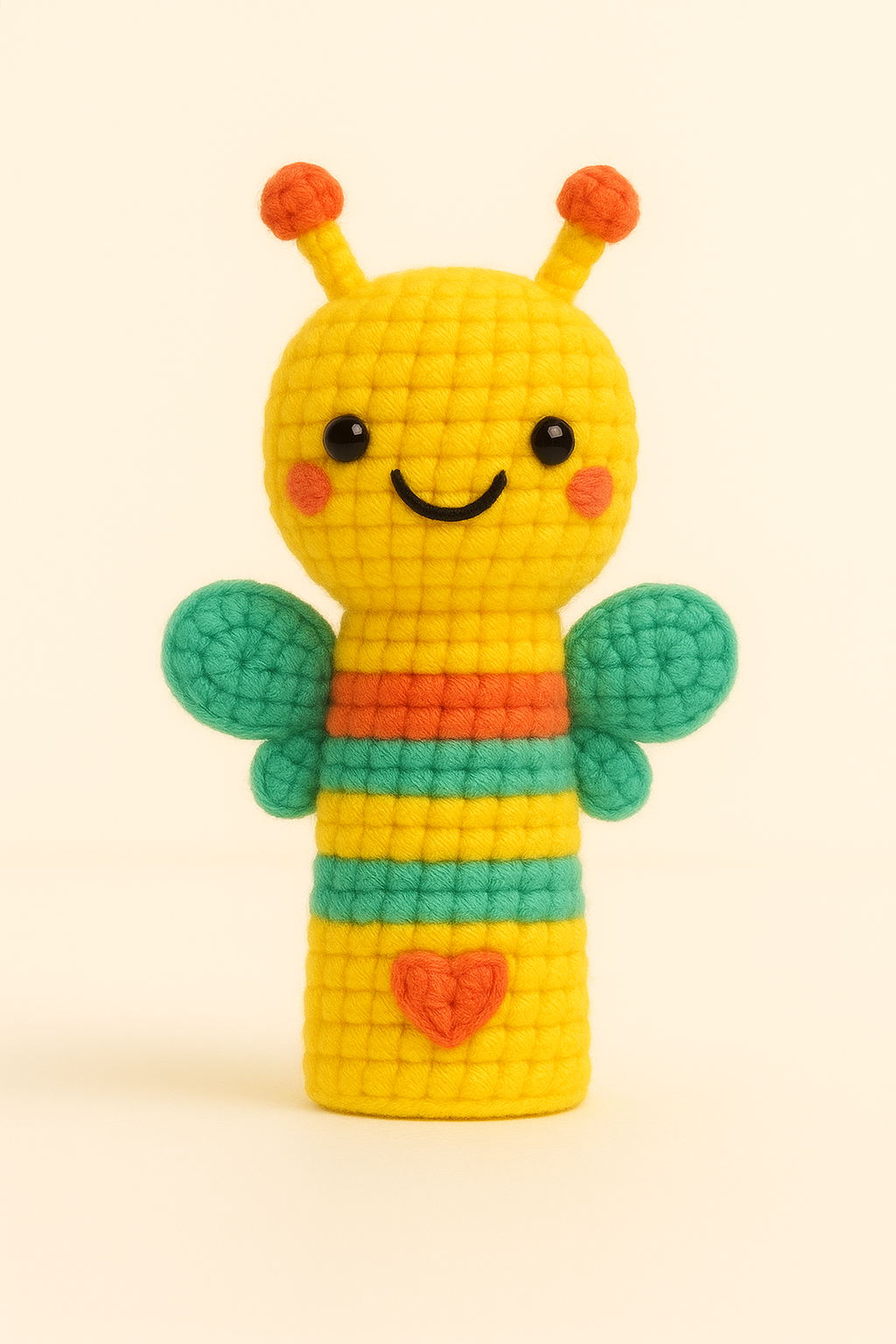 Colorful knitted plush toy resembling a cheerful bee with a yellow body, orange antennae, green wings, a red heart on its belly, and a smiling face with black eyes and a curved mouth.