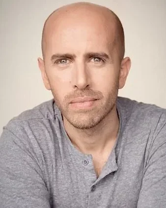 A man with a shaved head and light-colored eyes wearing a gray shirt, looking directly at the camera.