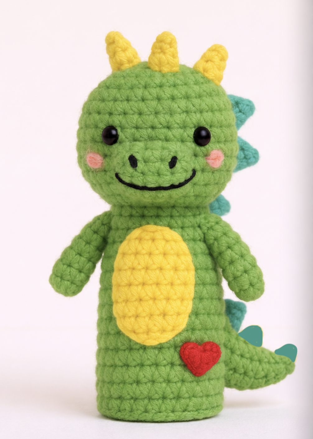 A crocheted plush toy resembling a cute green dinosaur with yellow horns, a yellow belly, and a small yellow heart on its lower body. The dinosaur has black eyes, pink cheeks, and a smiling face.