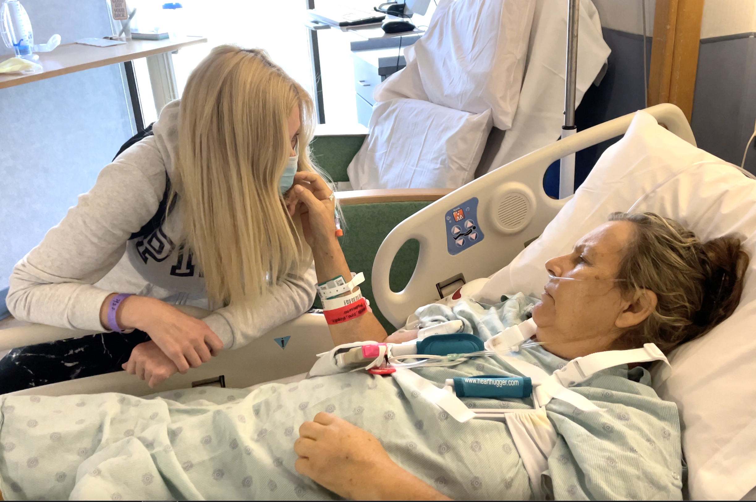 A woman with long blonde hair leans over a hospital bed, holding the hand of an older woman lying in the bed. The older woman has a nasal oxygen tube and various medical devices attached. Both women are touching foreheads, and the scene appears emotional and caring.