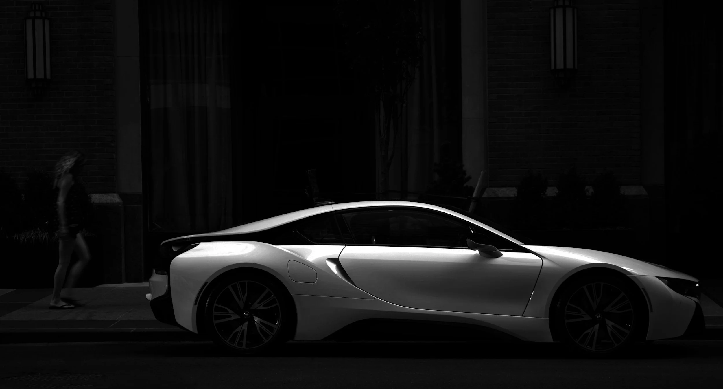 A sleek, modern sports car parked on a city street at night with a pedestrian walking nearby.