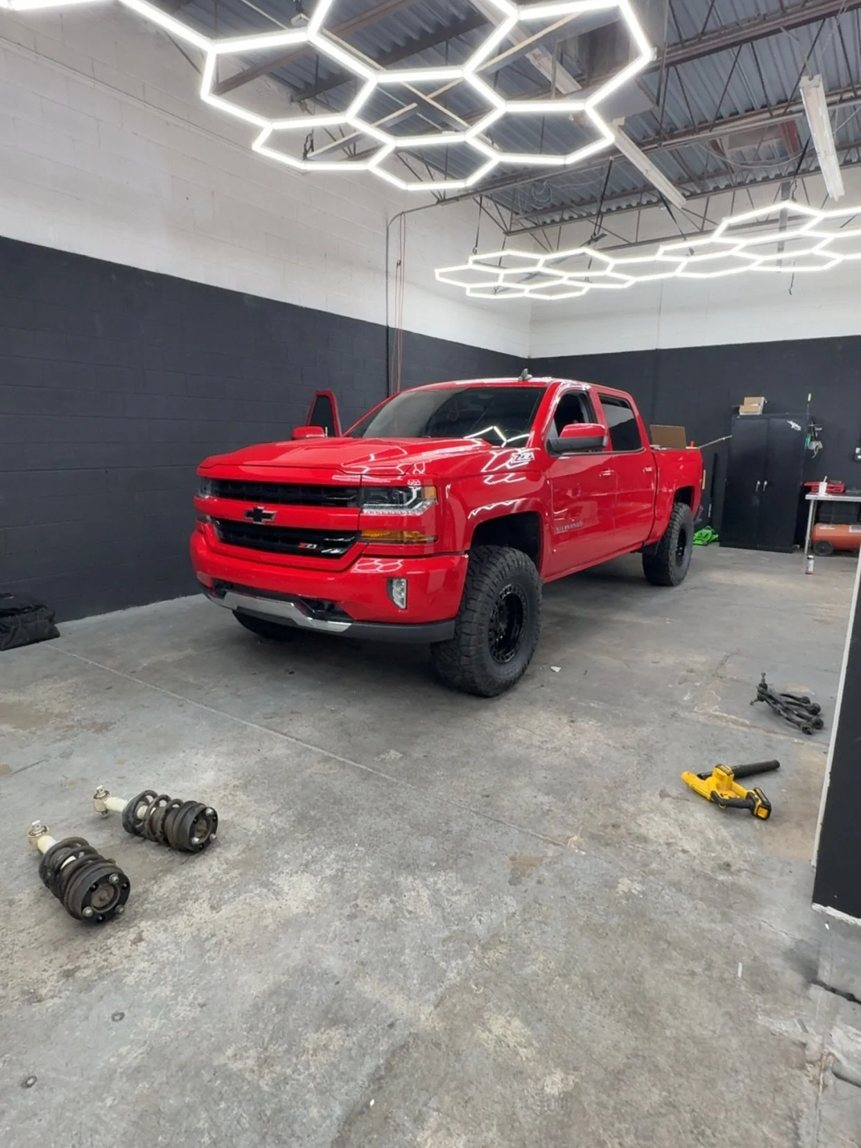 Red Chevrolet Silverado truck inside a garage with tools and spare parts on the floor, black wall, and hexagon-shaped ceiling lights.