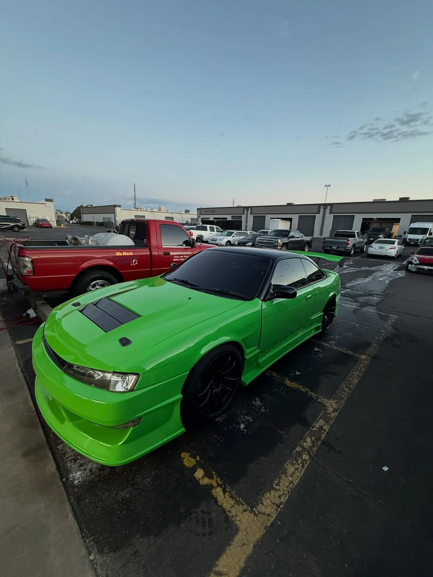A bright green sports car with black wheels and a black hood vent parked in a parking lot under a cloudy sky.